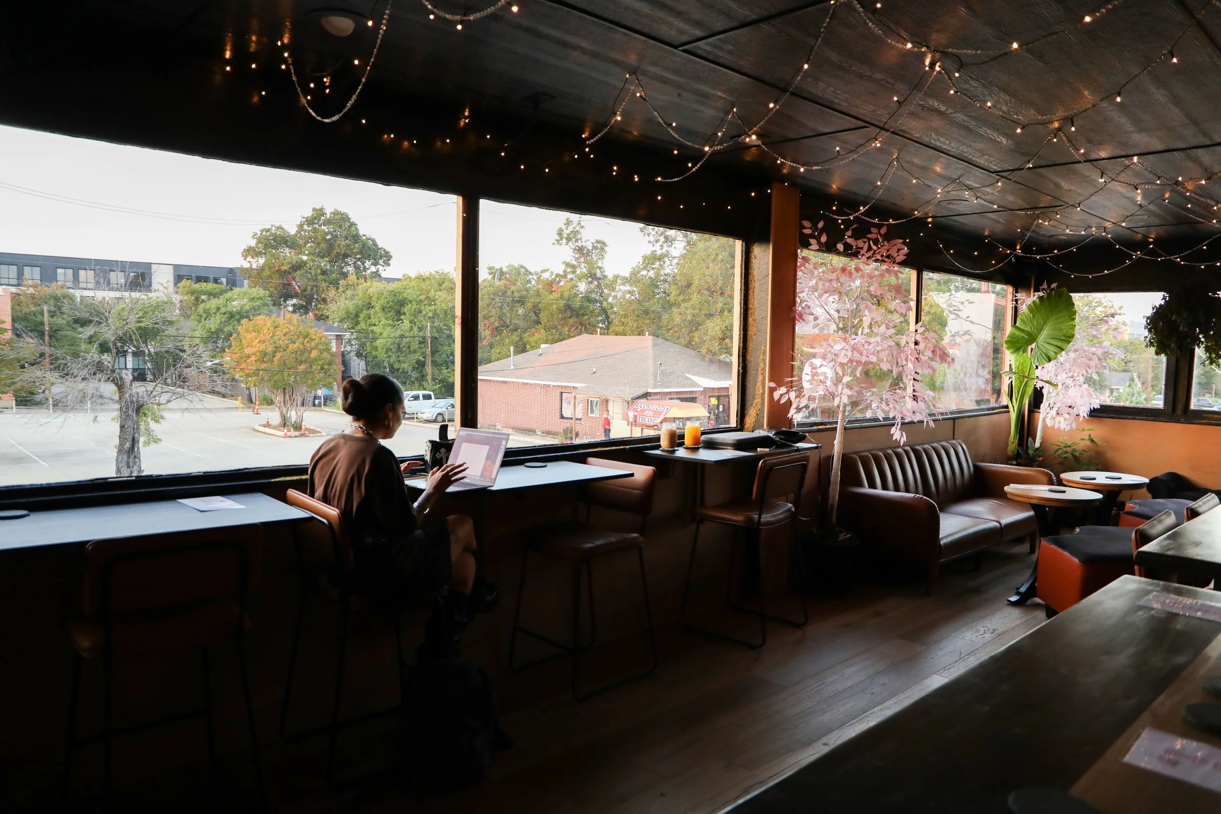 Interior of a cozy cafe with large windows overlooking trees and a parking lot, decorated with string lights and pink artificial trees, with a woman working on a laptop.