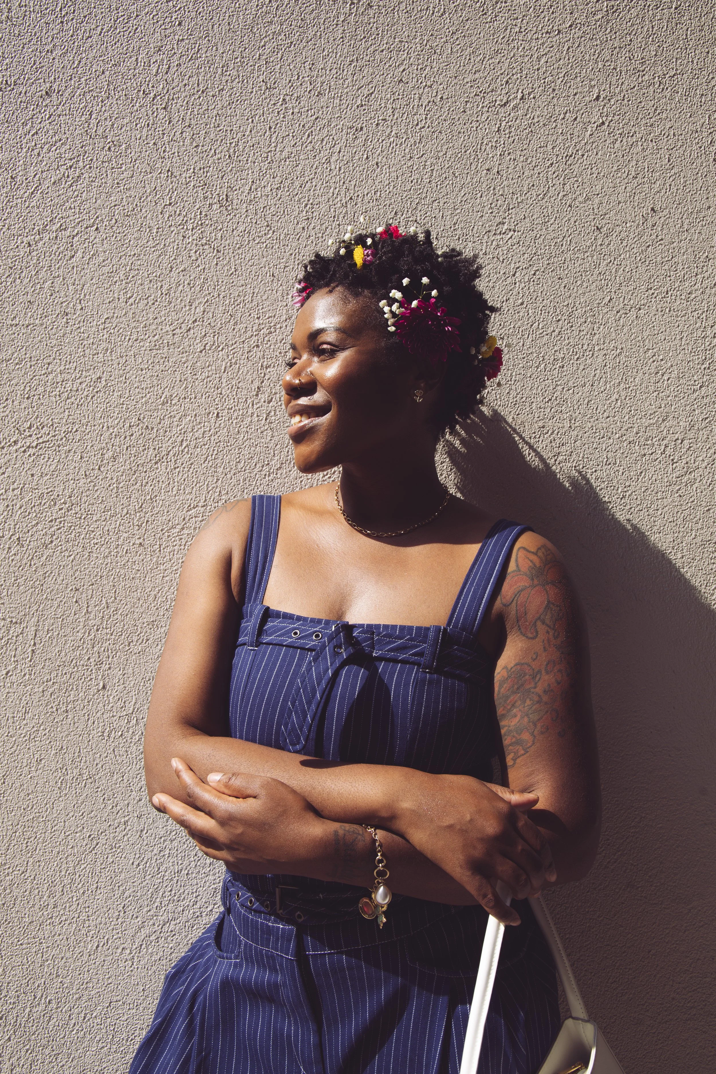 Local Dallas, Texas, DFW Non-traditional Fashion Editorial Creative Portraits Cover Art Photographer Photoshoot. @Afrodega . A woman with dark skin and short, curly hair decorated with colorful flowers, smiling and standing against a textured wall.