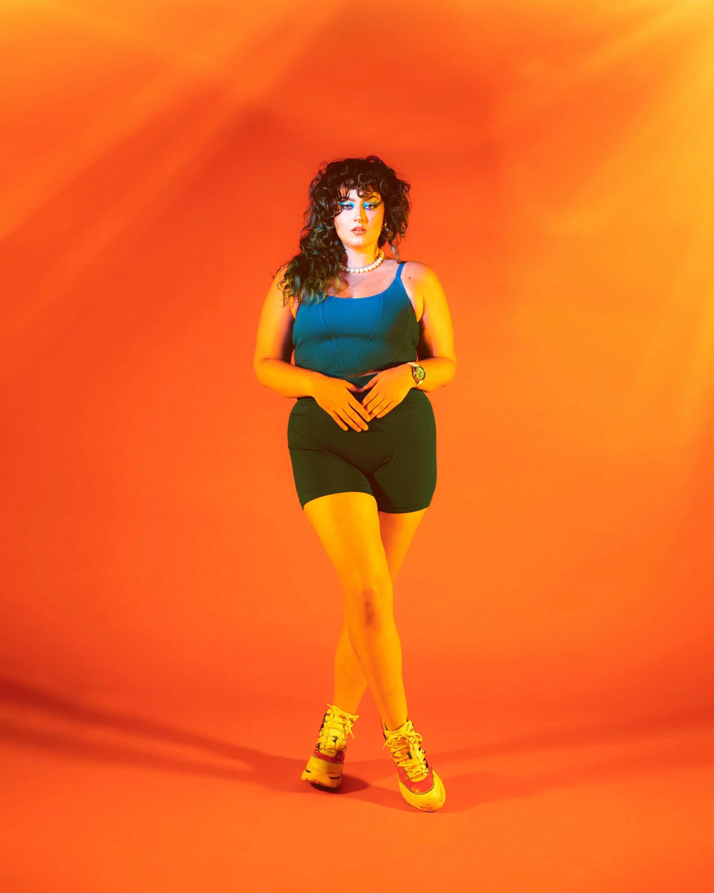 Local Dallas, Texas, DFW Non-traditional Fashion Editorial Creative Portraits Cover Art Photographer Photoshoot. Musician RAVS . A woman with curly dark hair wearing a blue tank top, black shorts, white sneakers, a pearl necklace, and a watch, standi