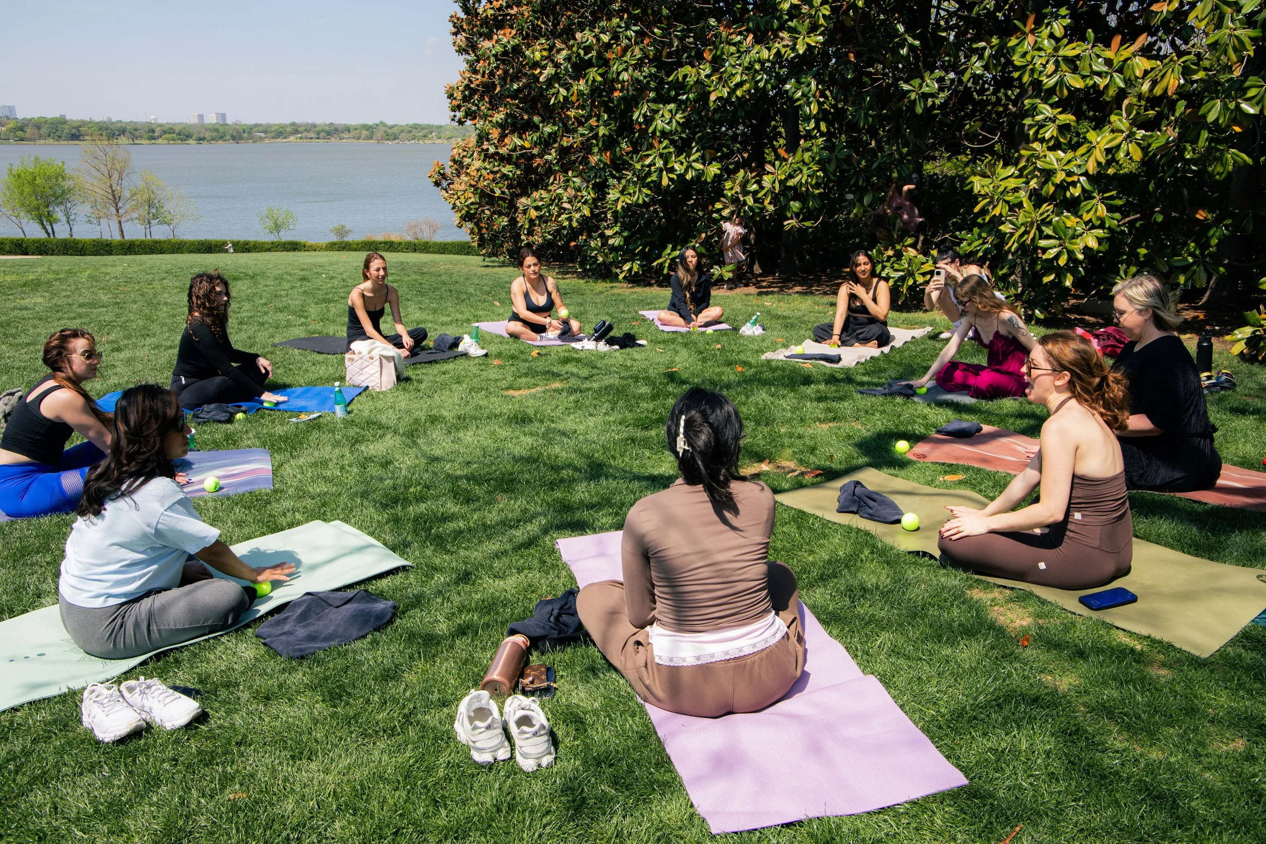 Group of women participating in a yoga class outdoors on a grassy park by a lake, with trees and a cityscape in the background.