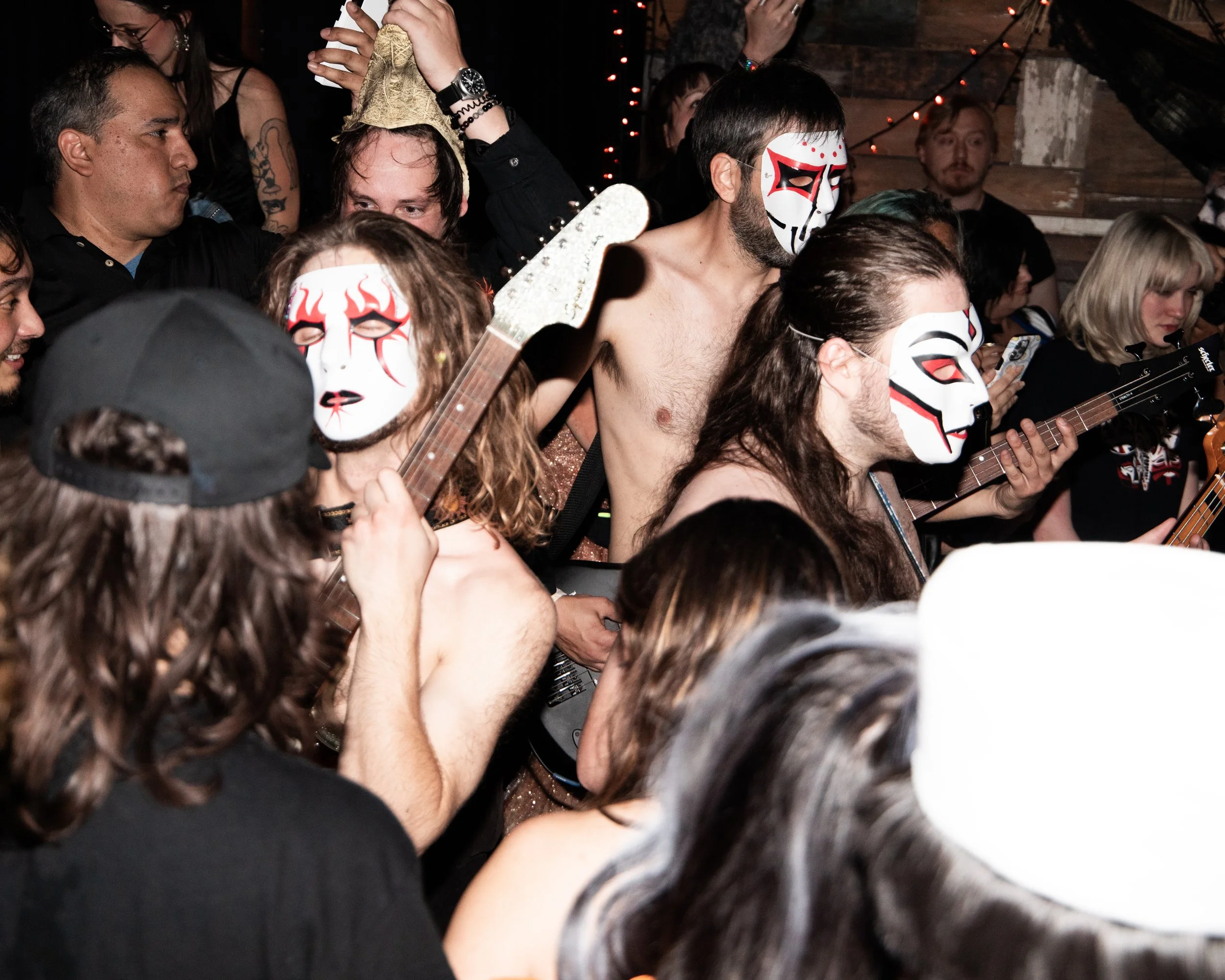 People at a lively party, some wearing masks, playing guitars, surrounded by a crowd in a cozy, dimly lit setting with wooden walls and string lights.