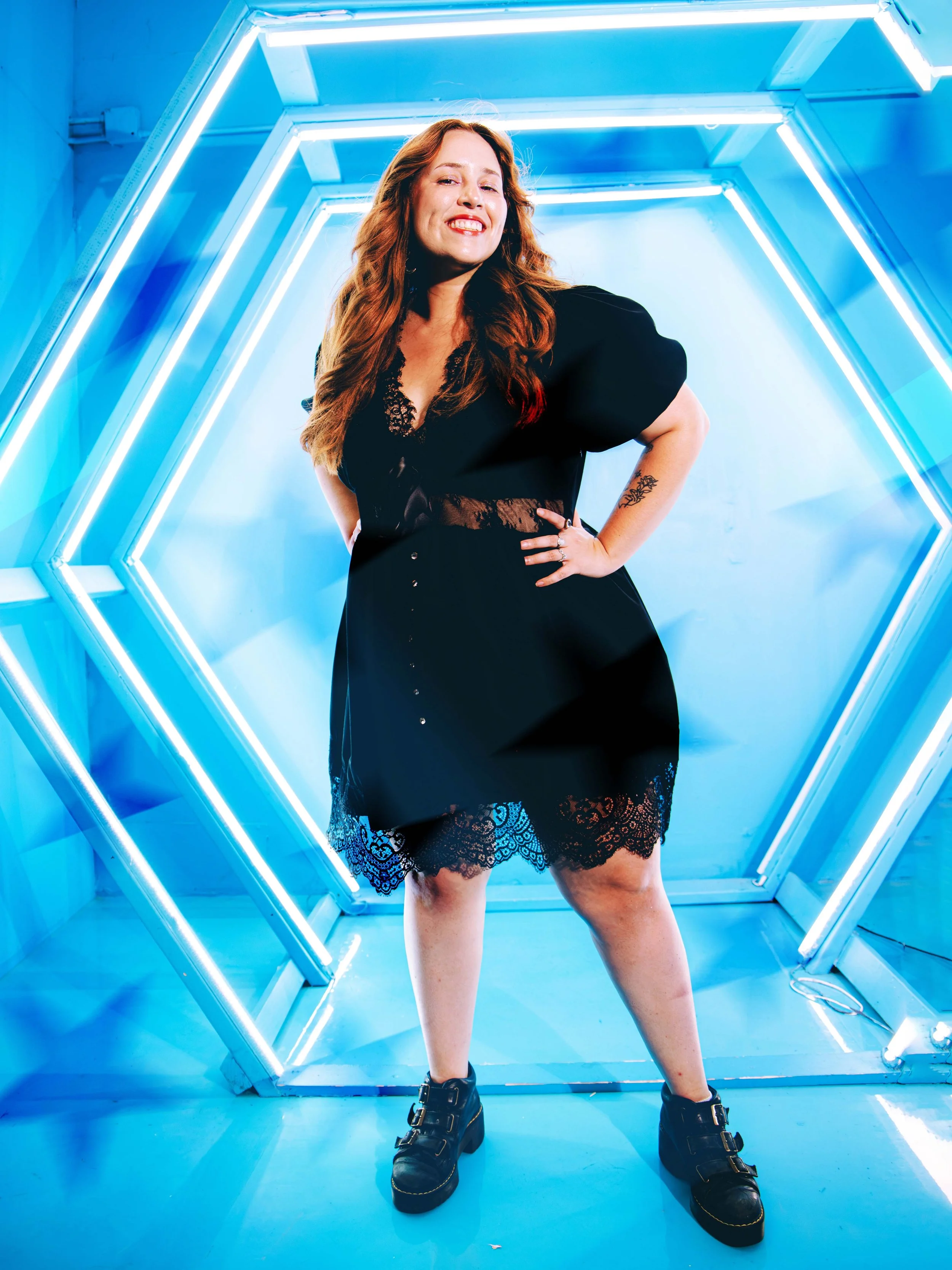 A woman with long red hair posing in front of a blue neon hexagonal tunnel, wearing a black dress with lace details and chunky black boots. Creative portrait.