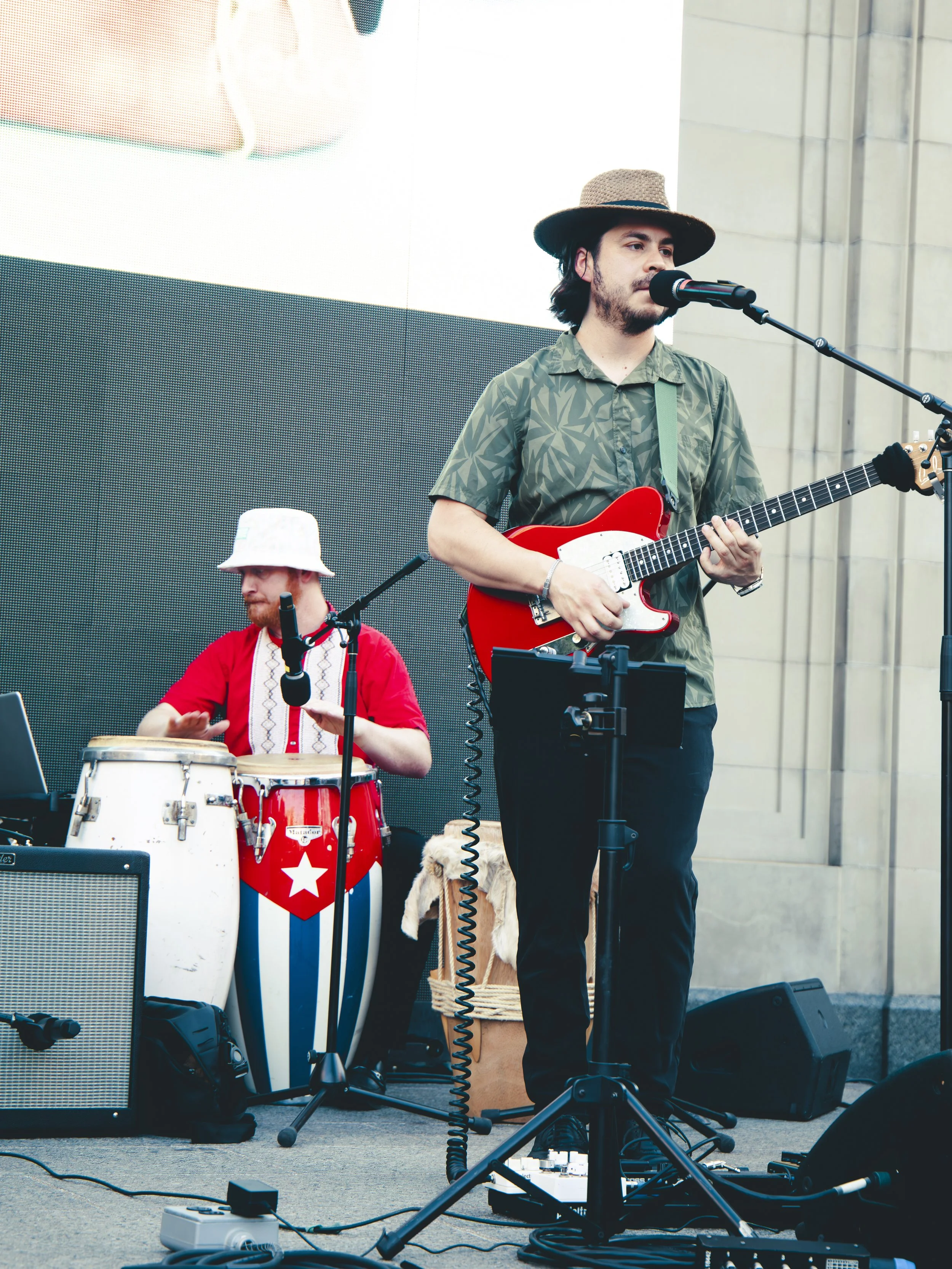 A male musician with long dark hair, wearing a straw hat and green patterned shirt, singing into a microphone while playing an electric guitar, with another musician in a red shirt and white hat playing percussion nearby, on an outdoor stage surround