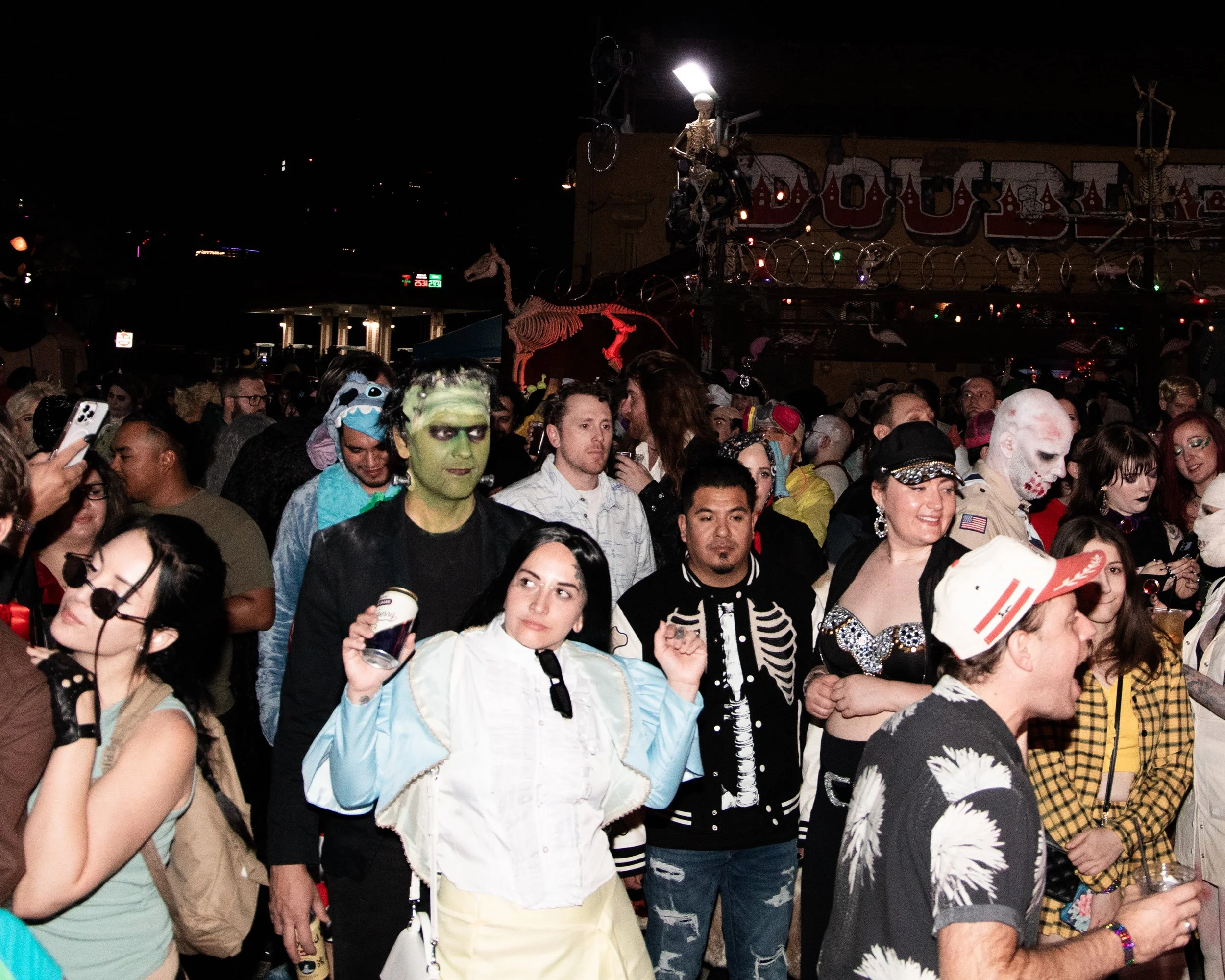 Crowd of people dressed in costumes at a nighttime event with a carnival or fair setting, including skeleton costumes, face paint, and accessories, with a booth decorated with skeletons and skeleton-themed decorations in the background.