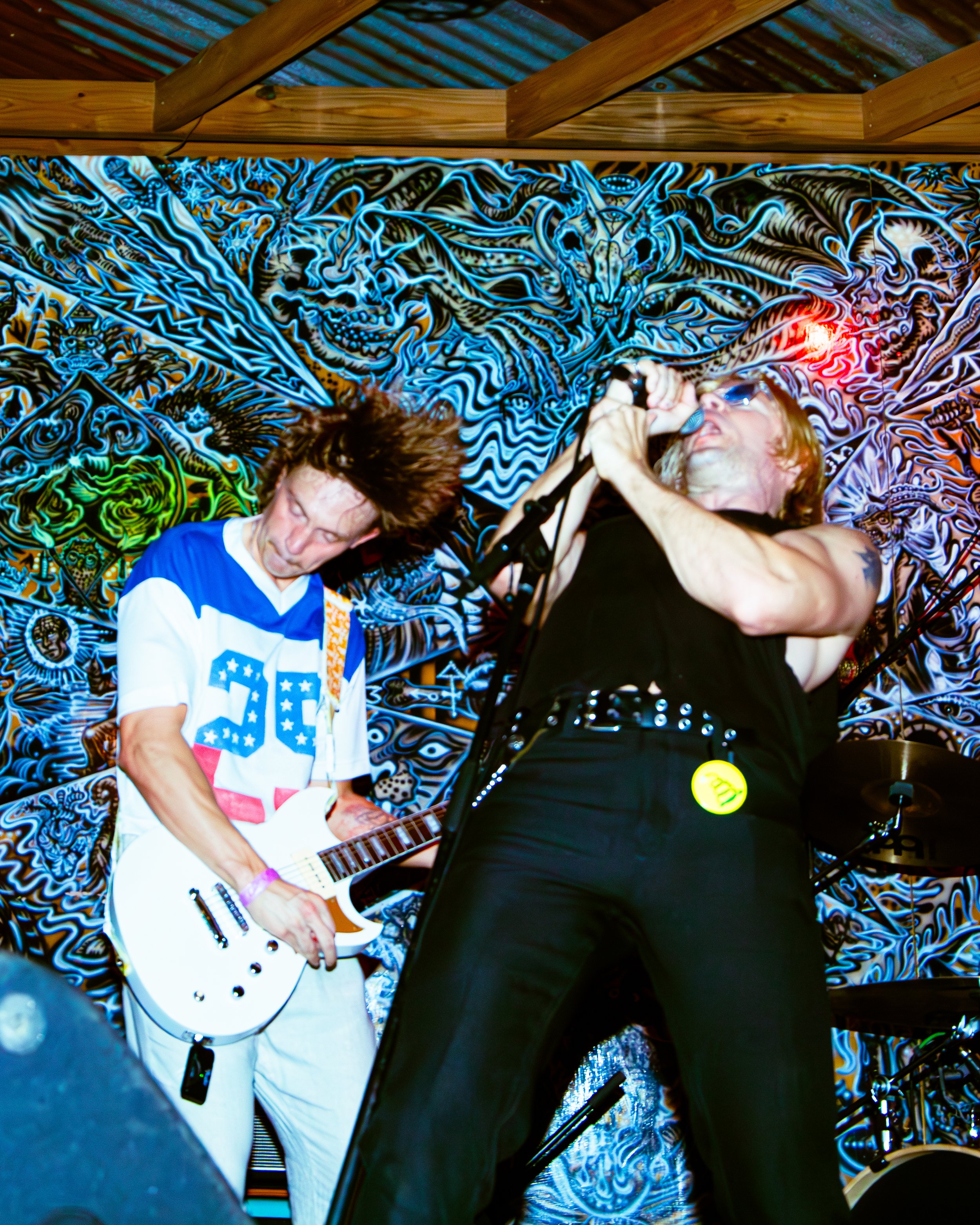 A musician with long hair playing an electric guitar, and another person singing into a microphone, in front of a vibrant, psychedelic background with intricate designs and patterns.