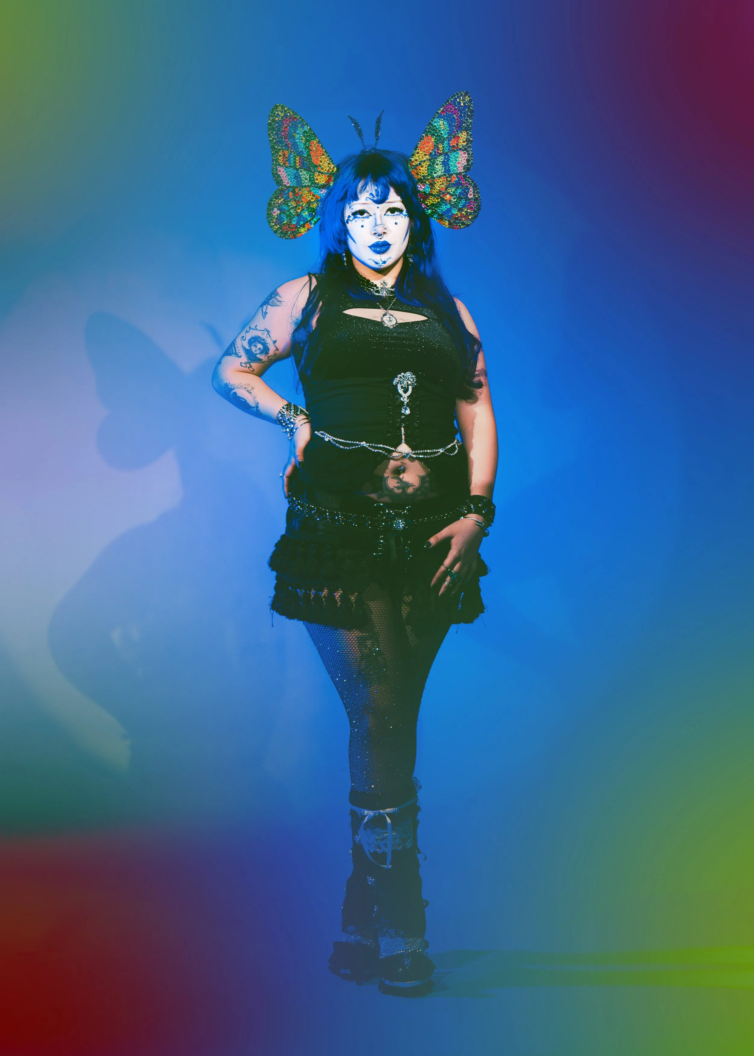 Local Dallas, Texas, DFW Non-traditional Fashion Editorial Creative Portraits Cover Art Photographer Photoshoot. Person with blue hair and butterfly headpiece, wearing black clothing, standing against a gradient blue background.