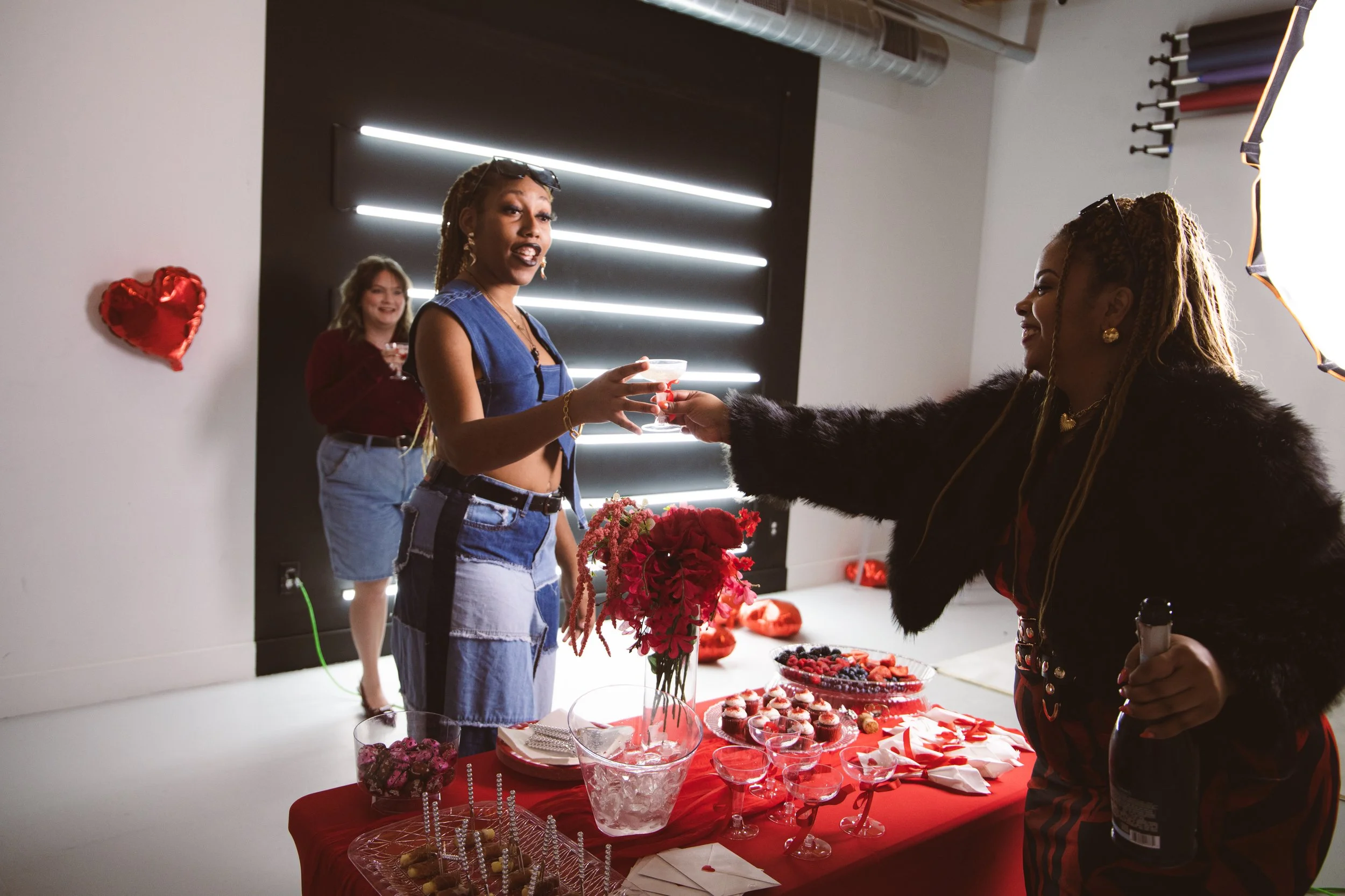 A woman with braids and hoop earrings receives a drink at a Valentine's Day celebration. Another woman in the background, with curly hair and a red top, smiles. A table with red flowers, cupcakes, candies, and other treats is in front of them, decora