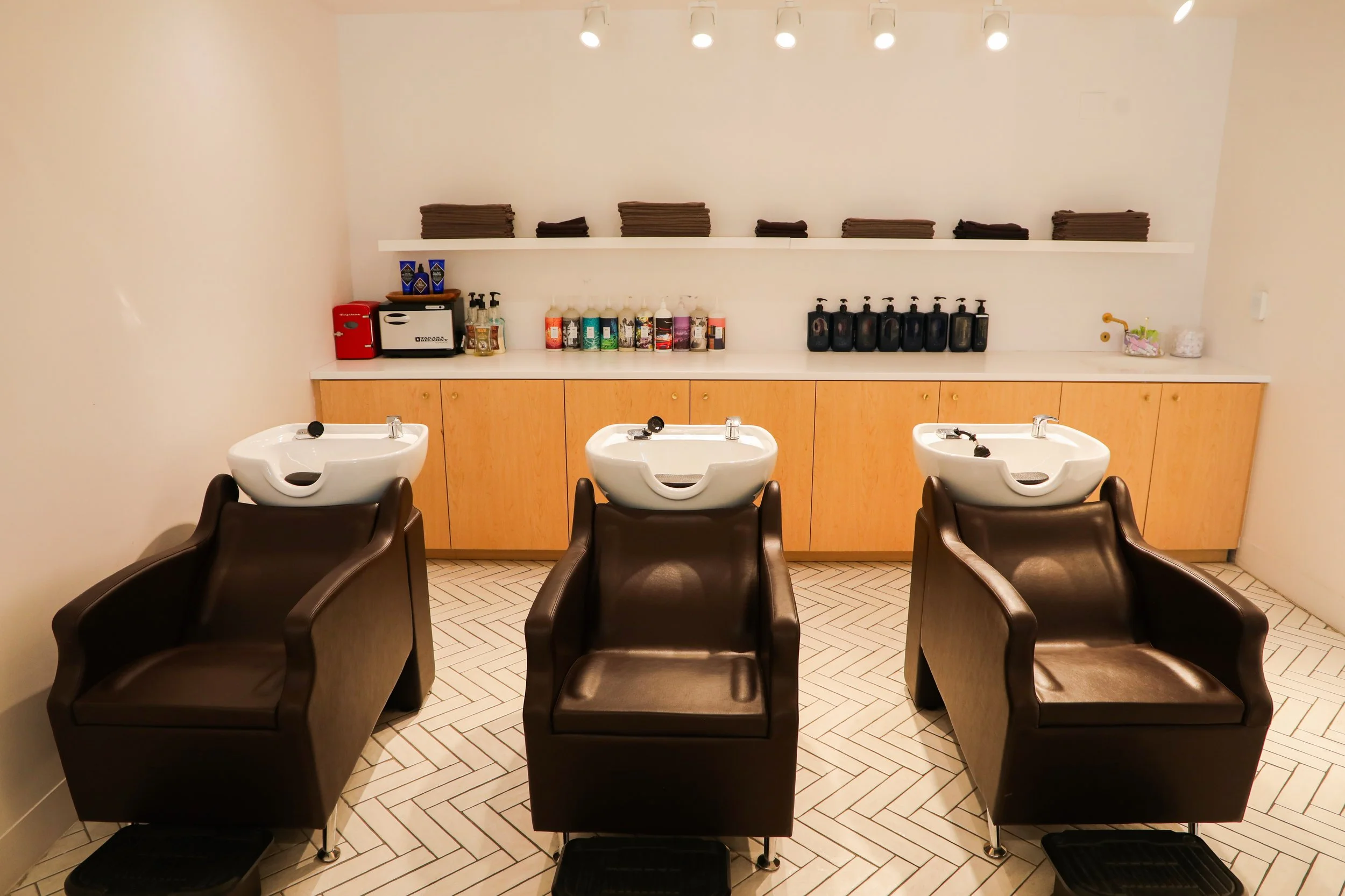 Salon with three black hair washing chairs in front of sinks, wooden cabinets, and shelves with folded towels and hair products.