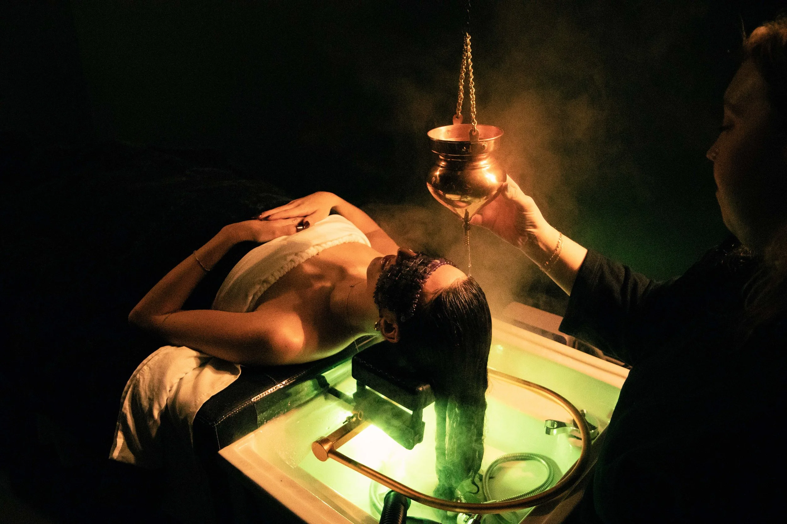 A person lying down on a bed with hands resting on their stomach, in a dark room illuminated by a small light. A priest or similar figure dressed in dark clothing is holding a metal chalice with a chain attached, positioned near the person's abdomen,