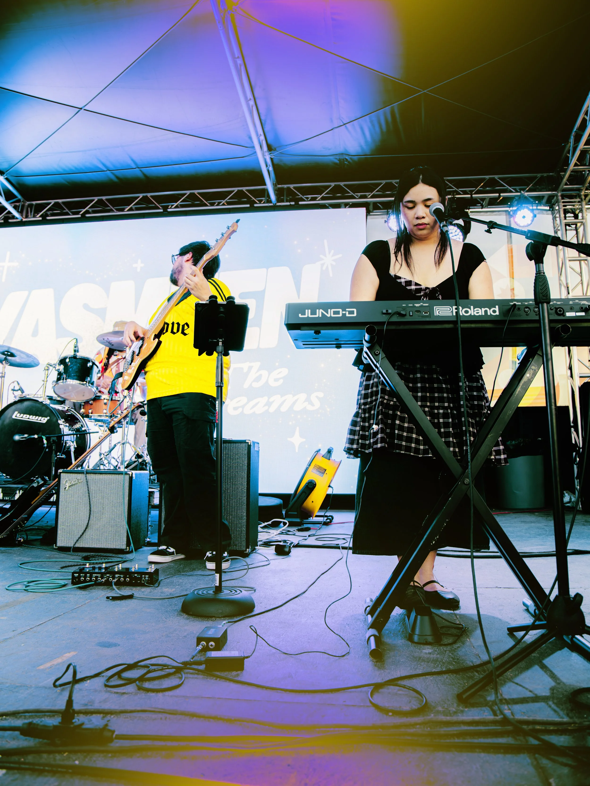 Performer playing keyboard on stage with backup band, large screen in background, stage lighting, and audio equipment.