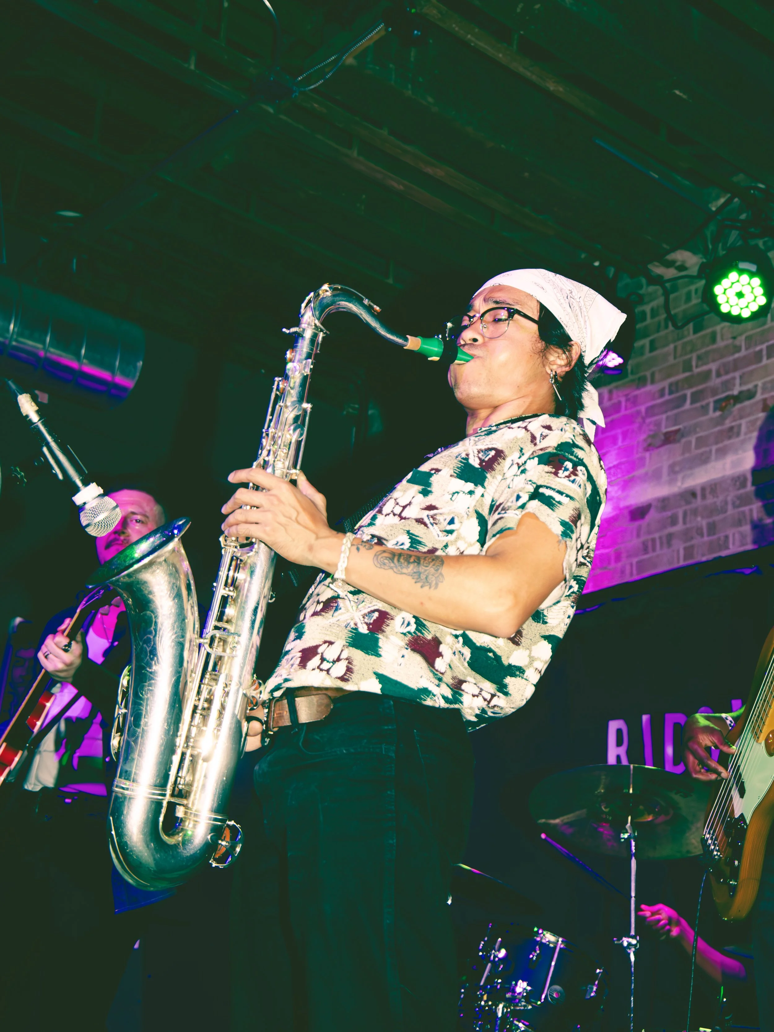 A woman in glasses and a white bandana playing a green mouthpiece saxophone on stage with colored lights and a brick wall background.