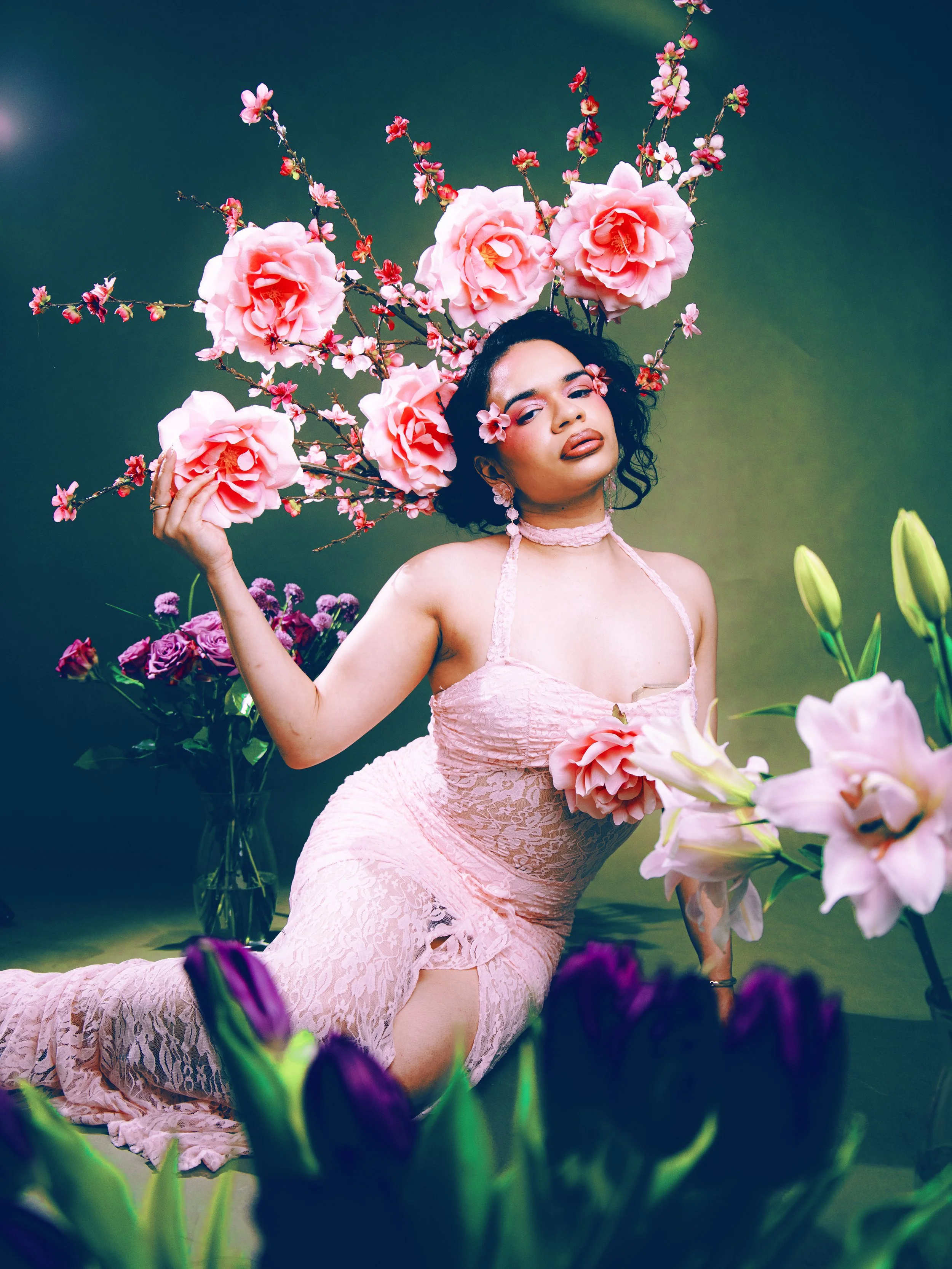 Local Dallas, Texas, DFW Non-traditional Fashion Editorial Creative Portraits Cover Art Photographer Photoshoot. Model @roseypears . A woman with dark curly hair and makeup, wearing a pink lace dress, poses among large pink flowers and smaller blosso