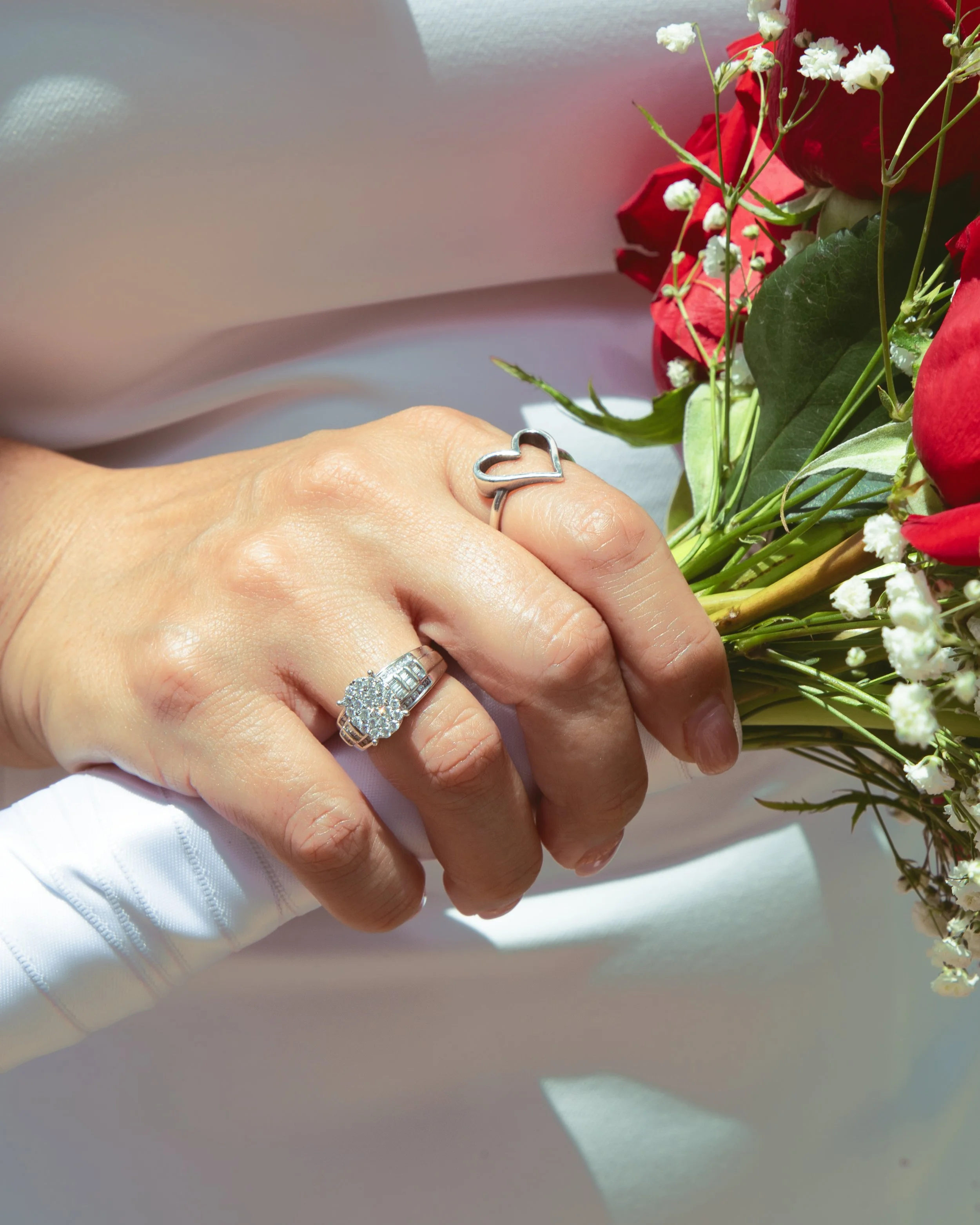 Close-up of a person's hand wearing a heart-shaped ring and a diamond ring, holding a bouquet of red roses and small white flowers, with a soft background.