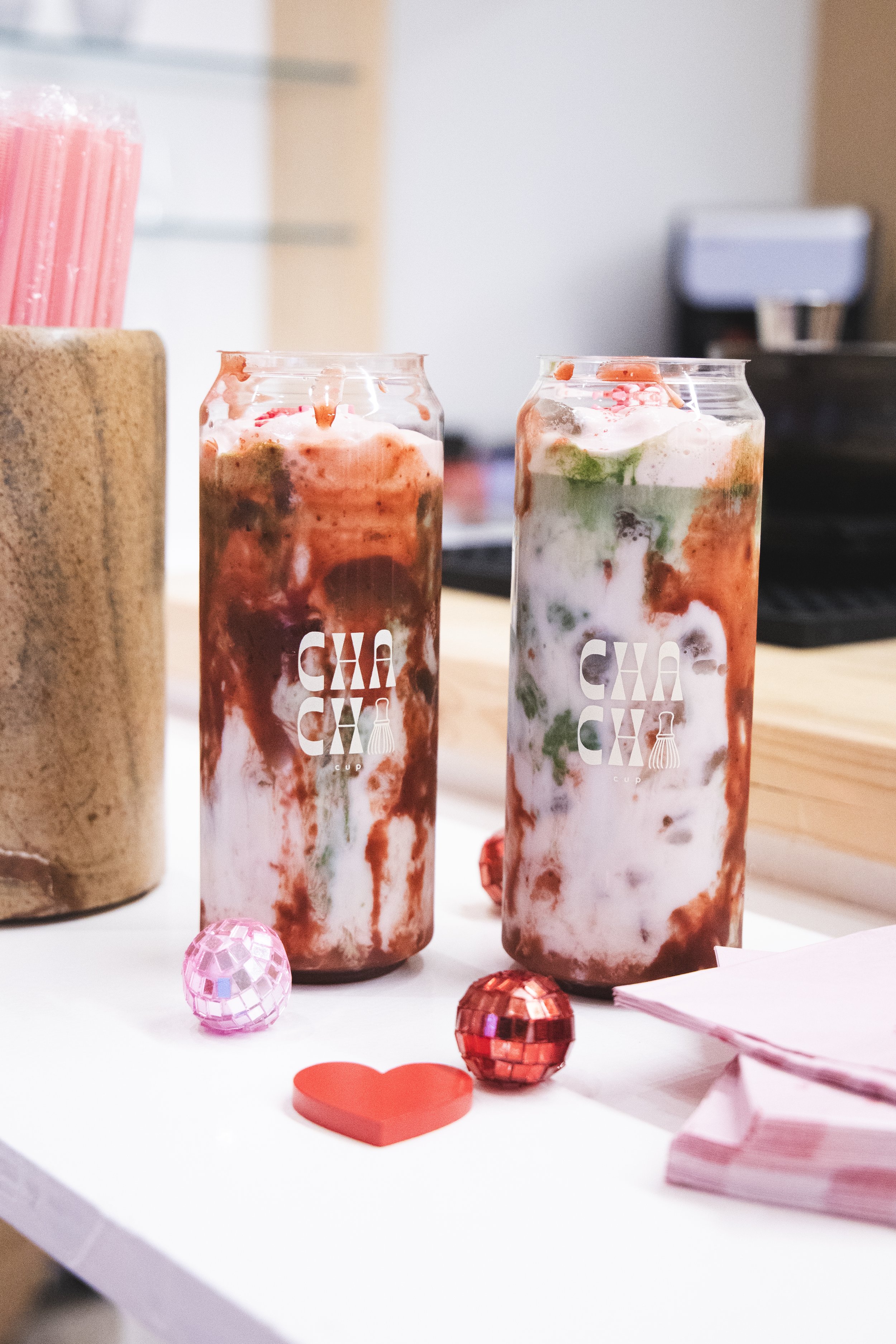 Two colorful smoothies in clear cups with the words CHACHACHA printed on them, surrounded by small decorative items including a pink globe, a red heart, and pink napkins.