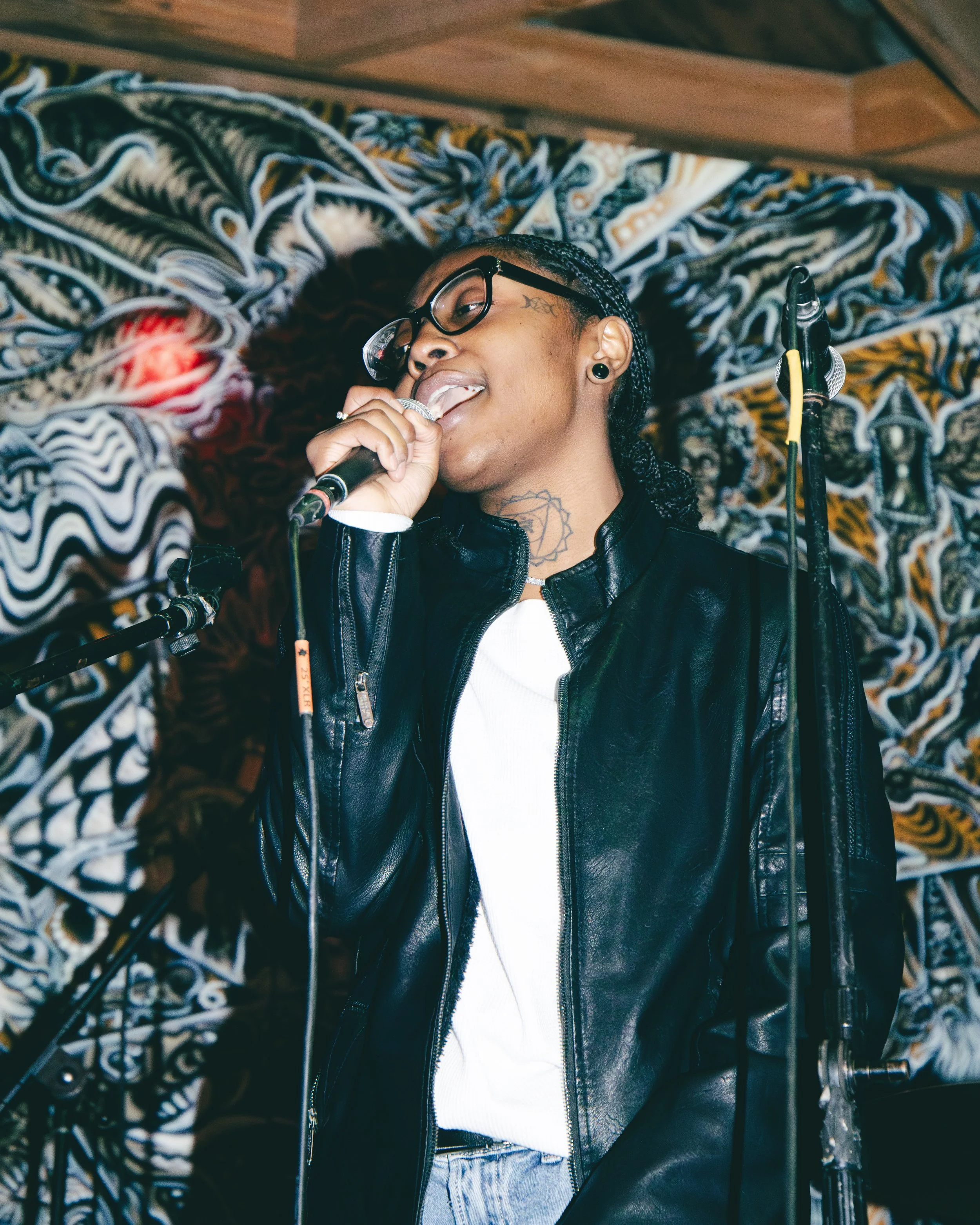 A person with short hair, glasses, and tattoos, singing into a microphone while wearing a leather jacket in front of a graffiti-covered wall.