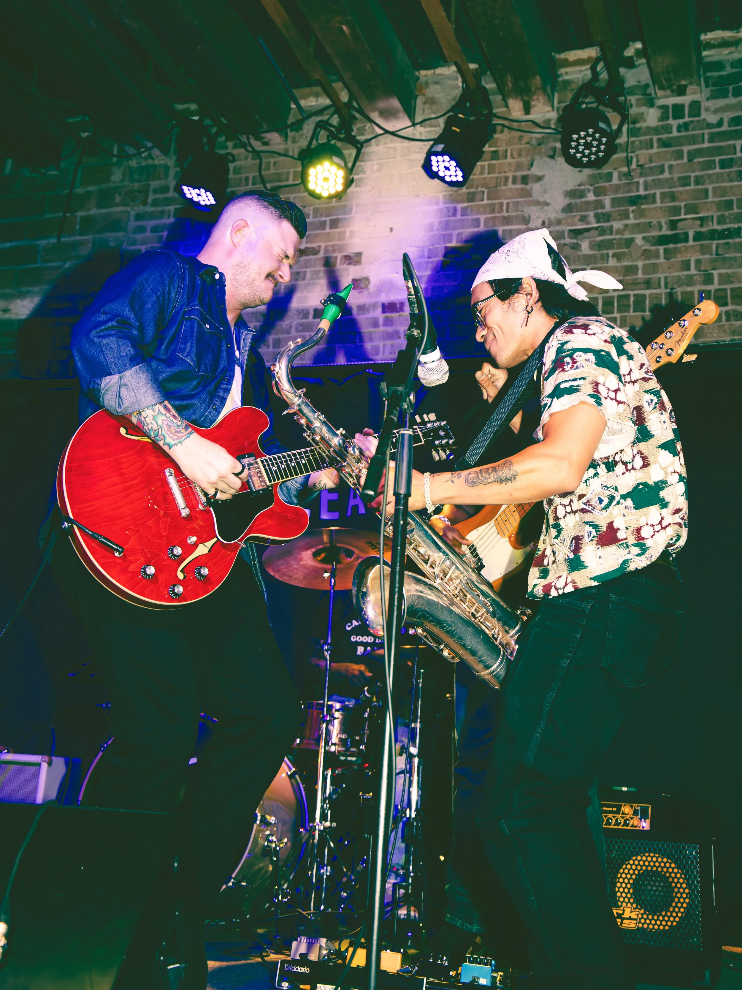 Two musicians performing on stage, playing guitars and drums, with colorful stage lights and brick wall background. Local Dallas, Texas musicians.