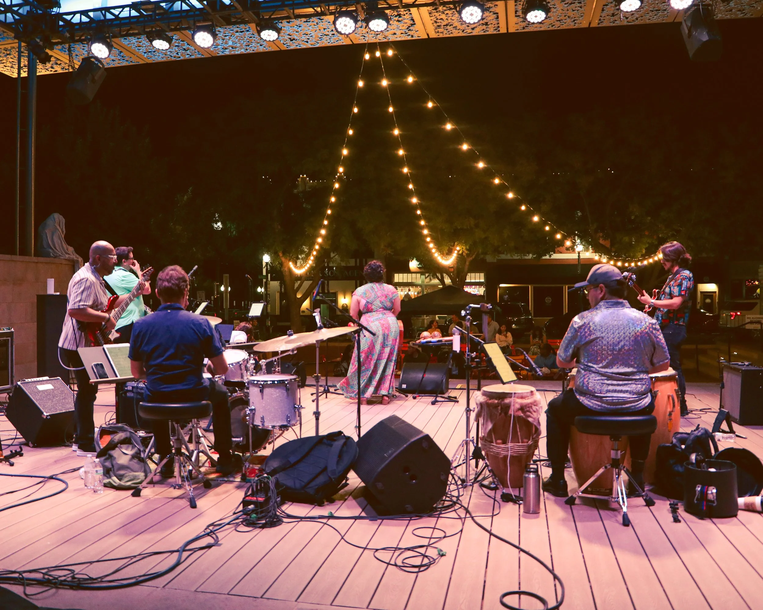 A group of musicians performing on an outdoor stage at night, with tables and audience in front, decorated with string lights hanging from the ceiling.