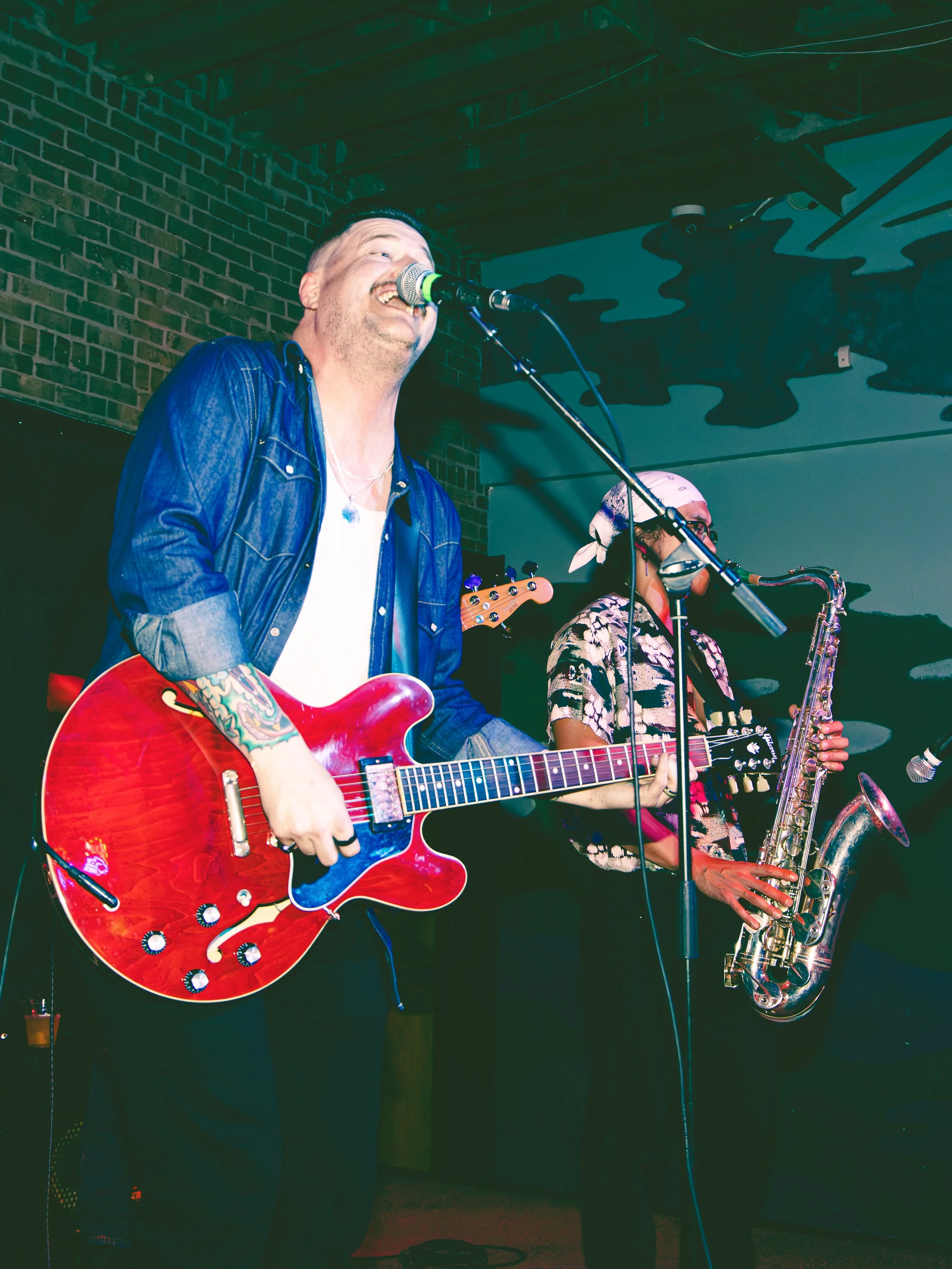 Musician singing into a microphone with an electric guitar, and a woman playing a saxophone in the background, in a dimly lit venue with brick walls and artistic ceiling design.