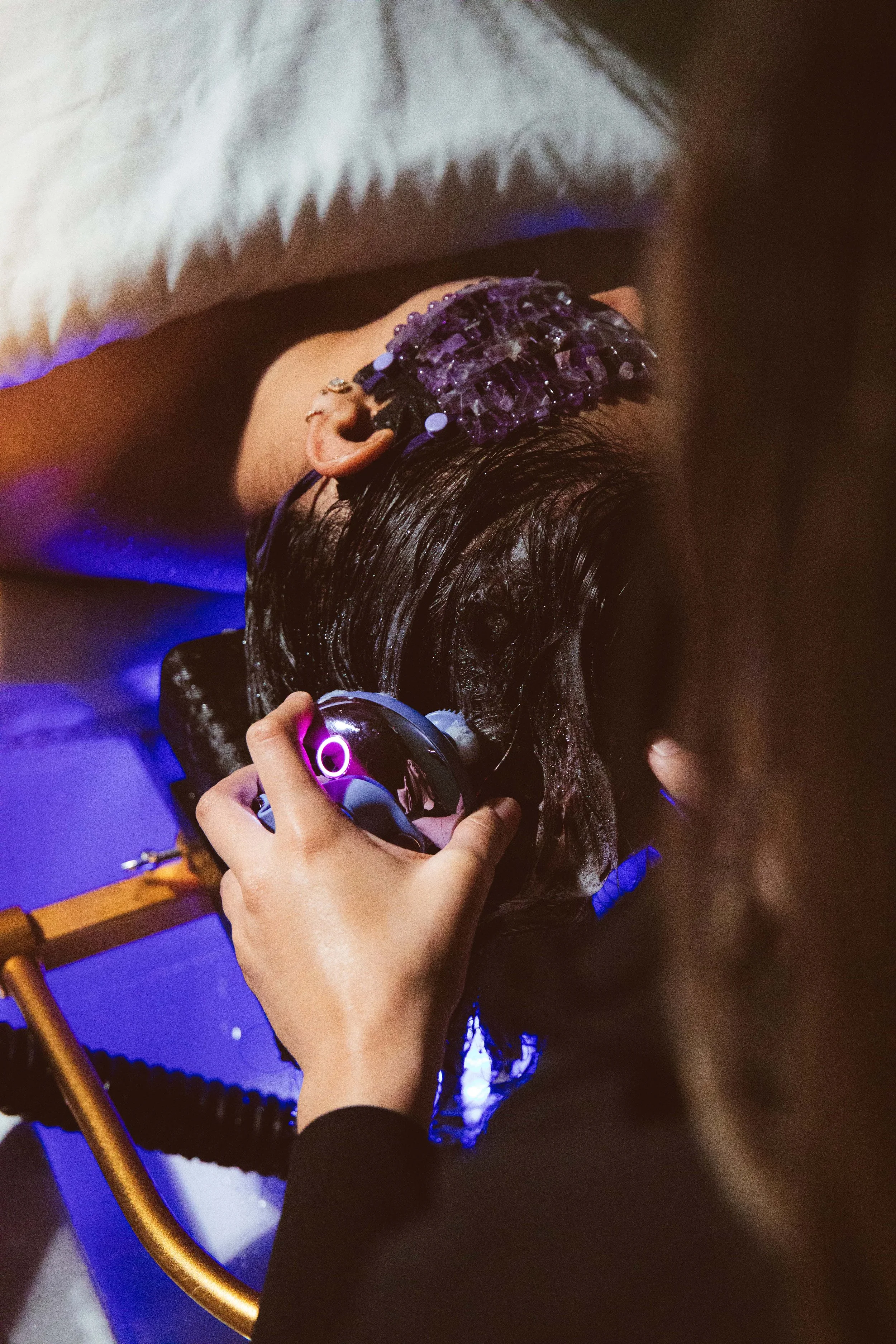 A person receiving a hair treatment with a purple crystal headpiece and a hair steamer device in a salon with purple lighting.