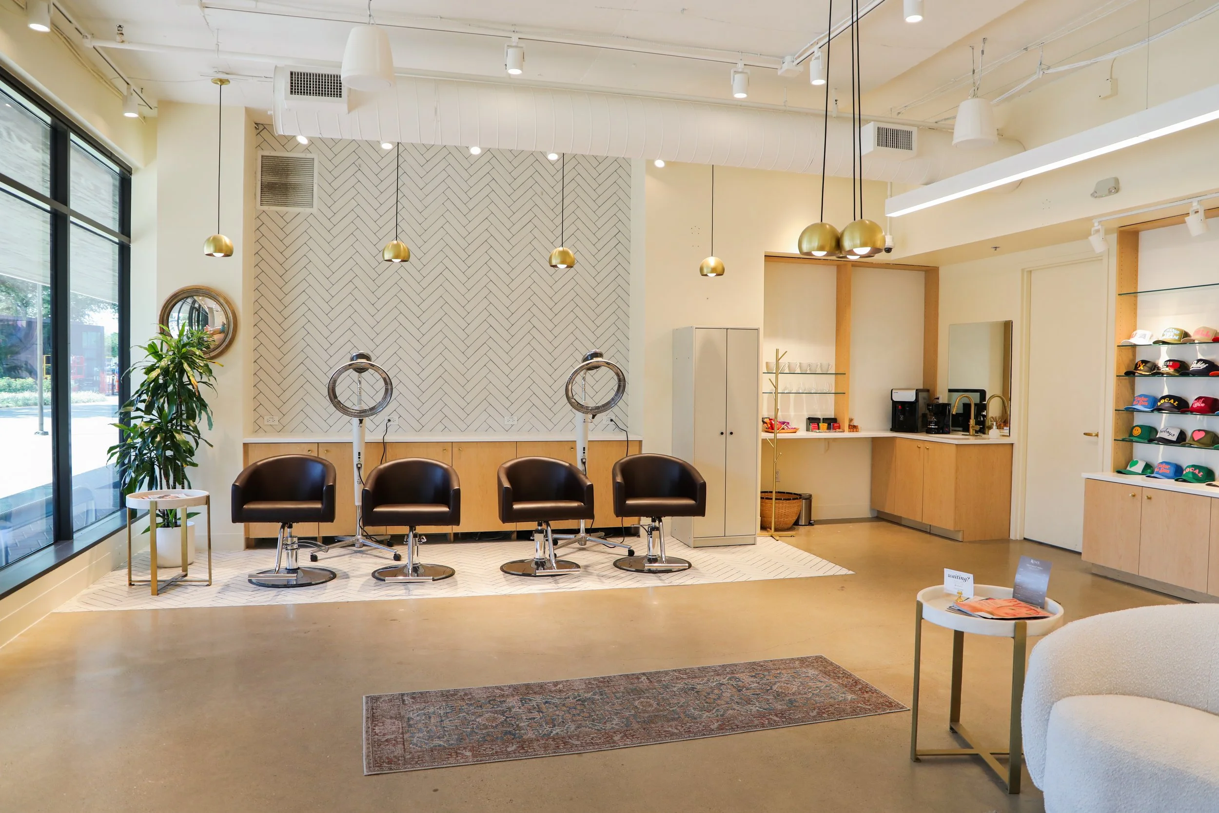 Interior of a modern hair salon with four salon chairs, hairdryers, and a large window on the left side. The wall has a herringbone tile pattern with hanging pendant lights, a plant, and a mirror. The right side features a counter with hats, glasses,