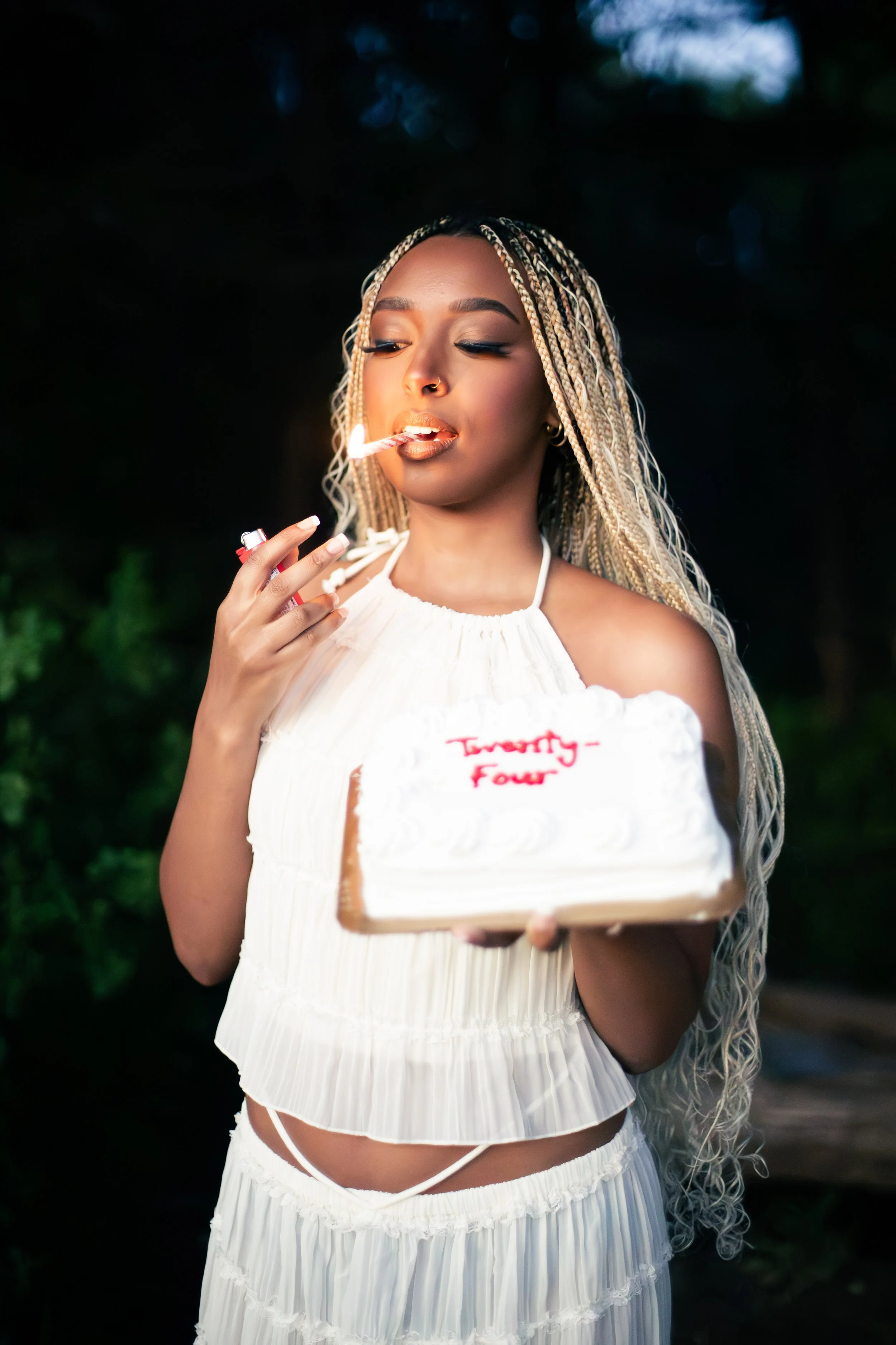 A woman with braided blonde hair and makeup, holding a birthday cake with lit candles, wearing a white ruffled top and matching skirt, standing outdoors at dusk.