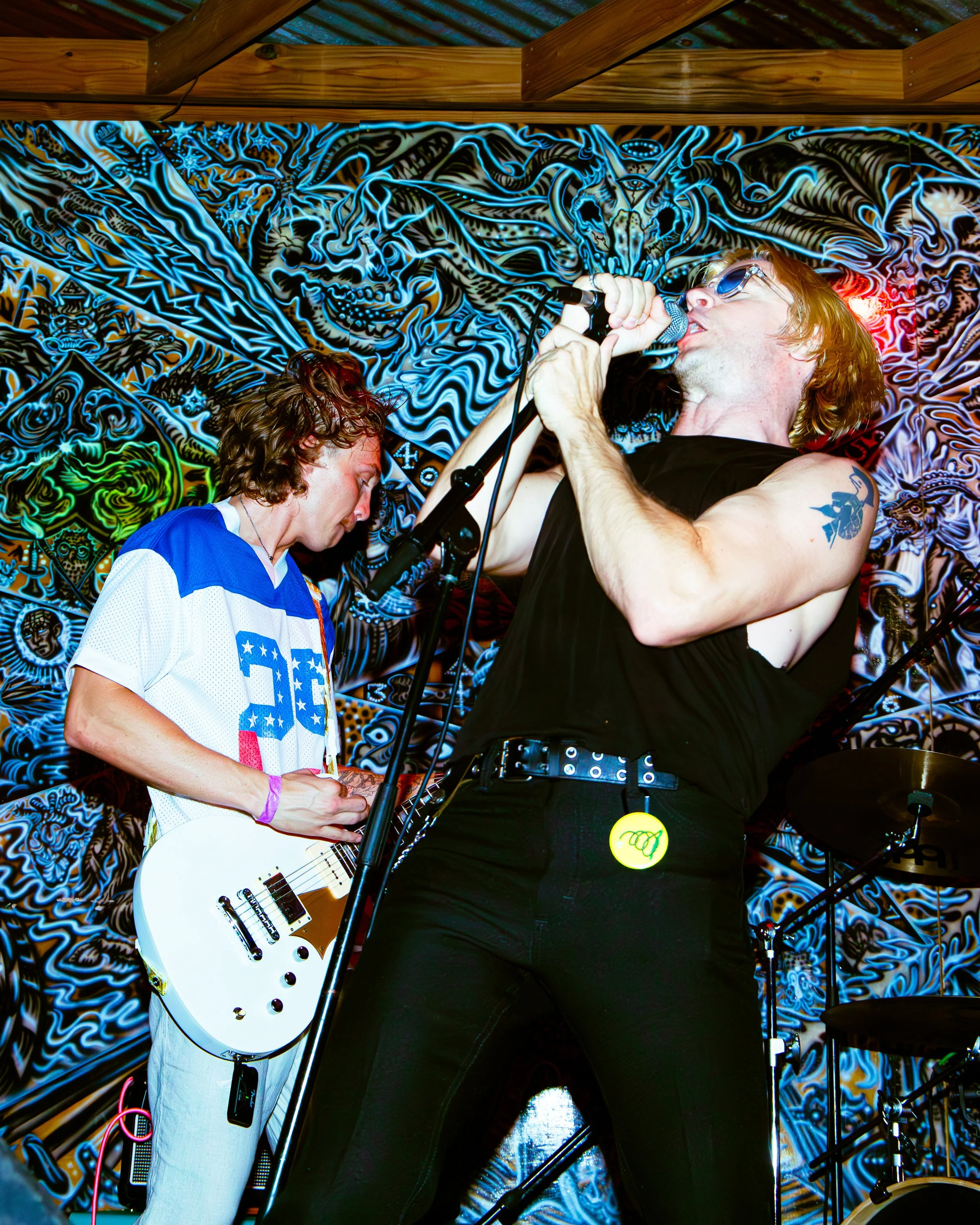 Two musicians perform on stage with a psychedelic and graffiti-inspired backdrop. One plays a white electric guitar, and the other sings into a microphone wearing sunglasses, with a tattoo on their arm.