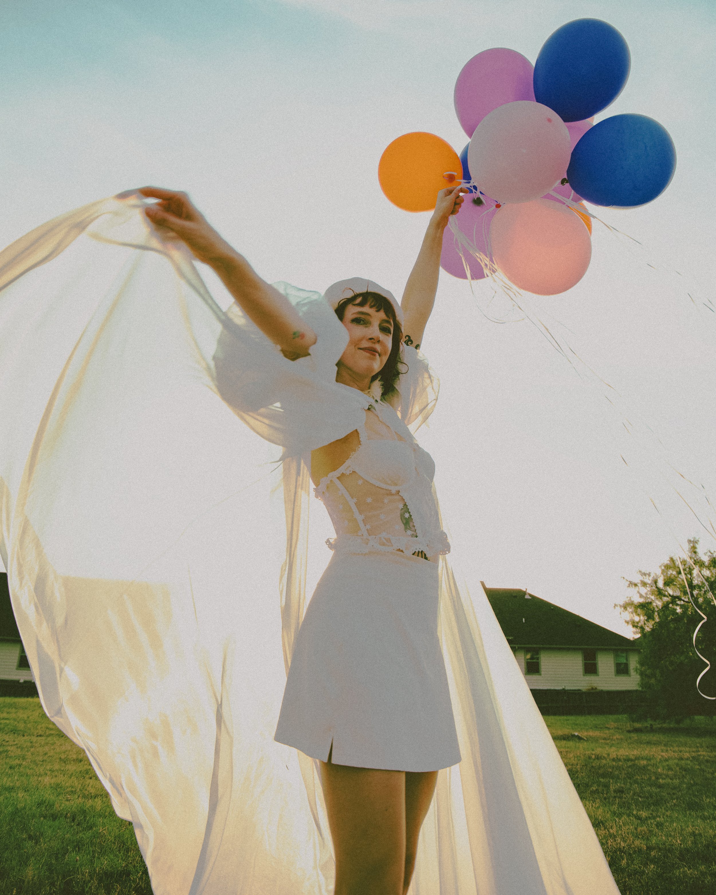 Local Dallas, Texas, DFW Non-traditional Fashion Editorial Creative Portraits Cover Art Photographer Photoshoot. A woman wearing a white dress and hat holds pink, purple, blue, and orange balloons outdoors on a sunny day, with a building and trees in