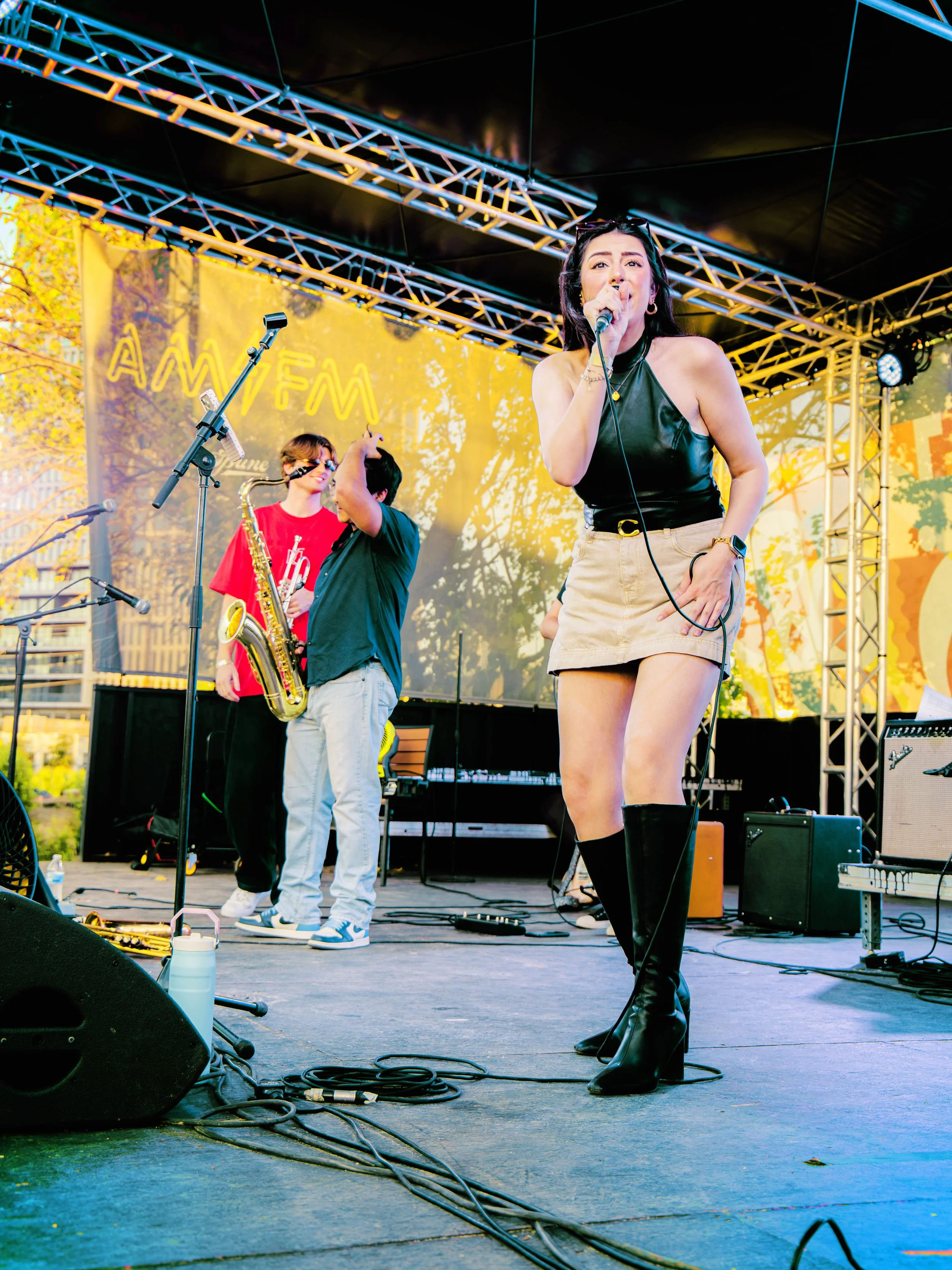 A woman singing into a microphone on an outdoor stage, with a yellow and orange backdrop and musicians with instruments in the background.