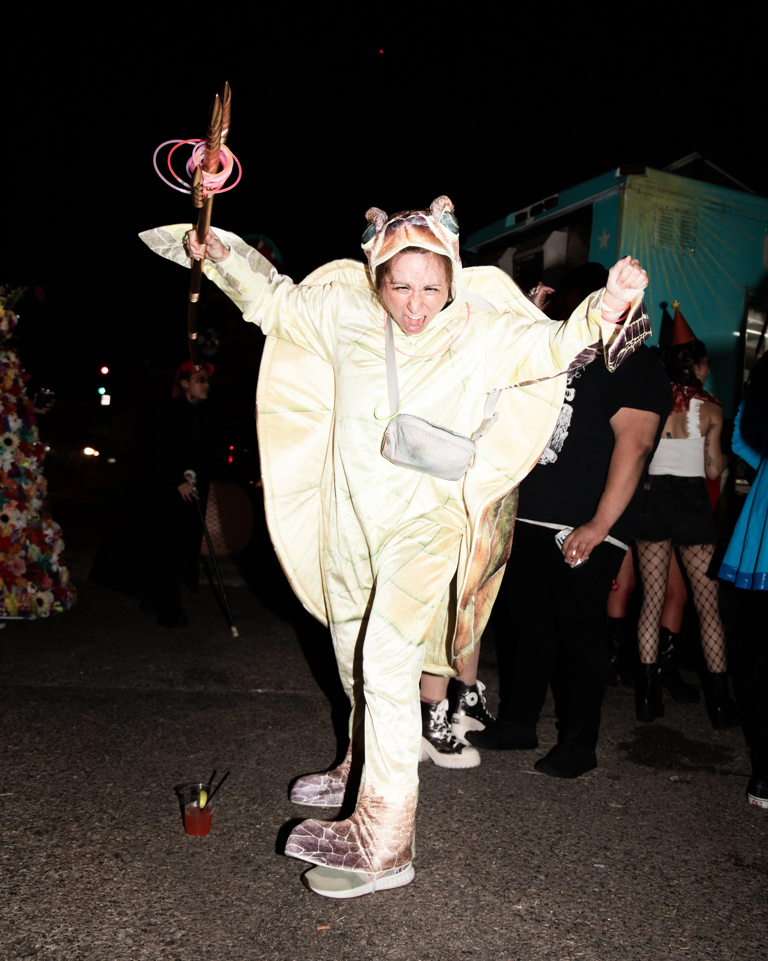 Person dressed in a giraffe costume with a hood, raising their arms and holding a staff with pink swirling lights at a nighttime event. Crowd of people in various costumes visible in the background, some wearing fishnet stockings and themed outfits.