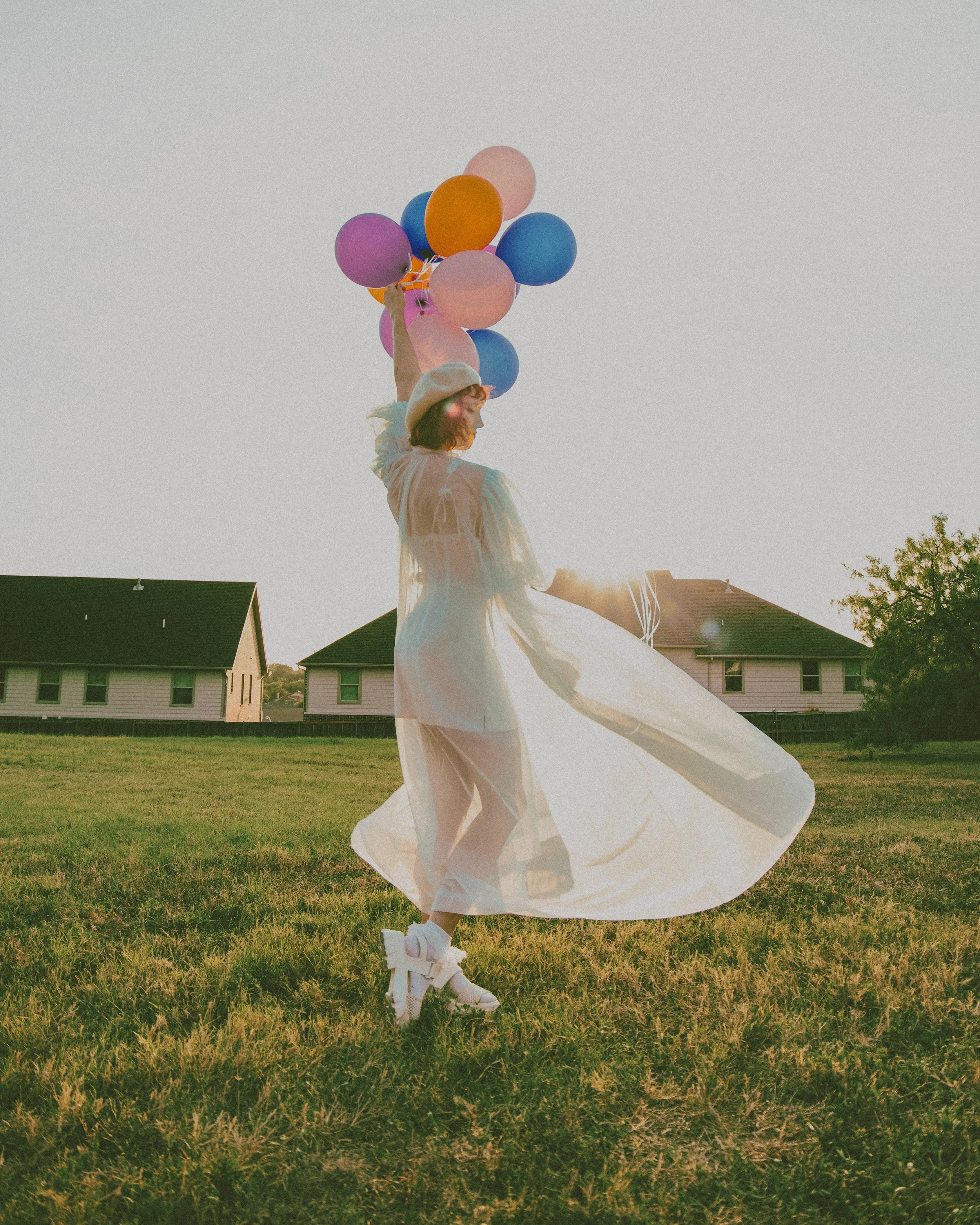 Woman standing outdoors in a grassy area holding colorful balloons with the sun setting behind her.