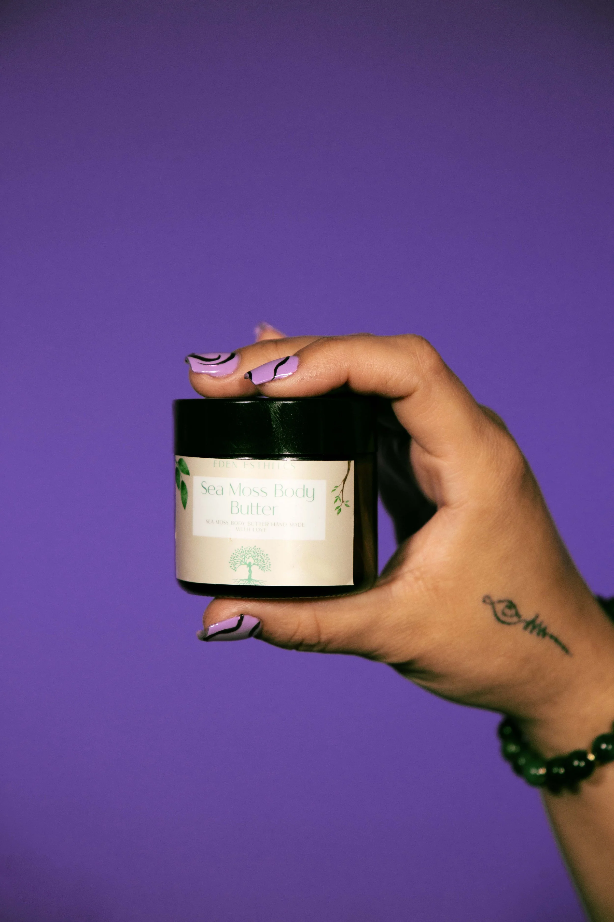 A hand holding a jar of sea moss body butter against a purple background. The person's nails are painted with lavender and black wavy designs, and they are wearing a green beaded bracelet and a tattoo of the word 'love' on the wrist.