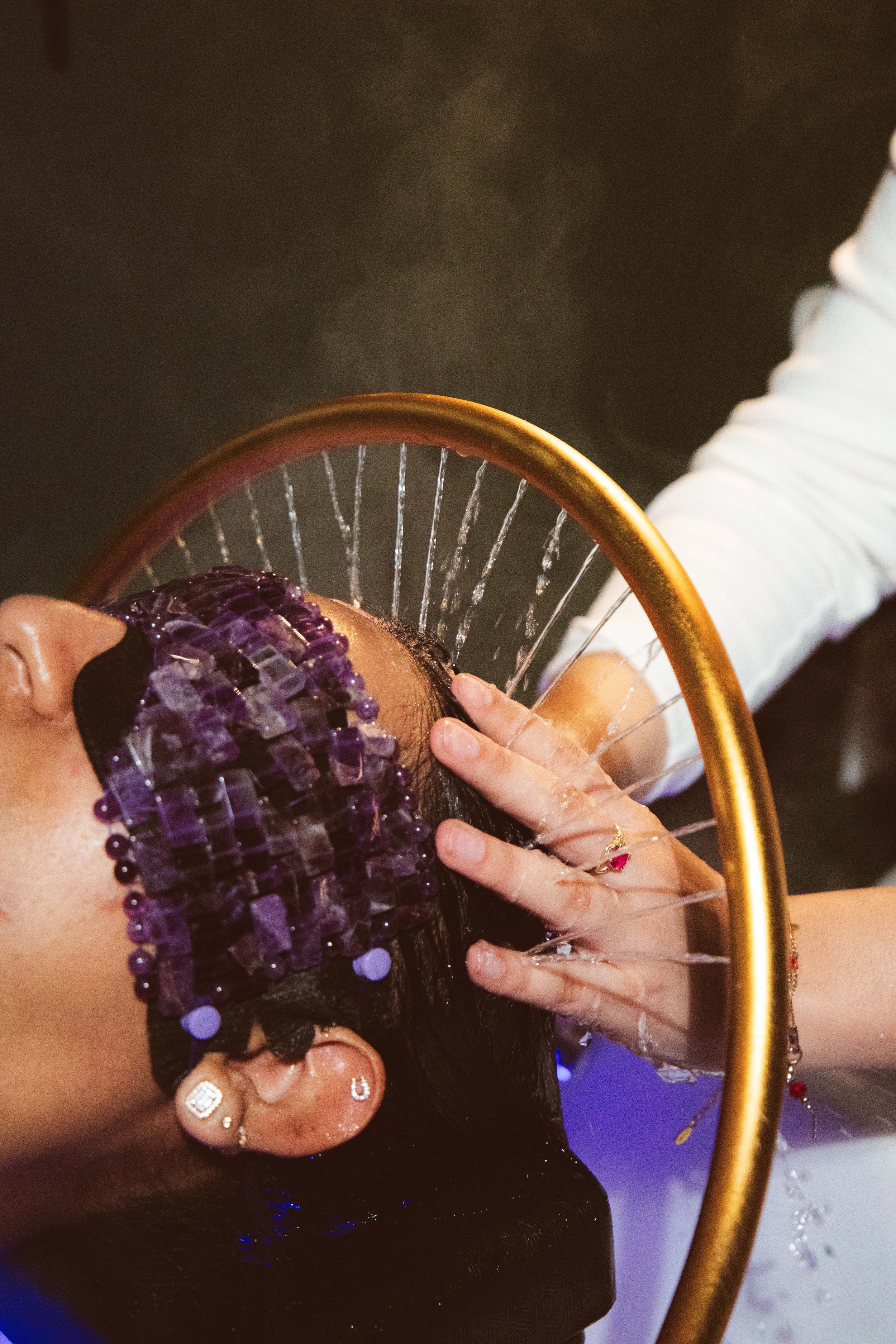 A person with dark hair, purple hair rollers, and jewelry receives a head massage with a curved instrument in a spa or beauty salon setting.