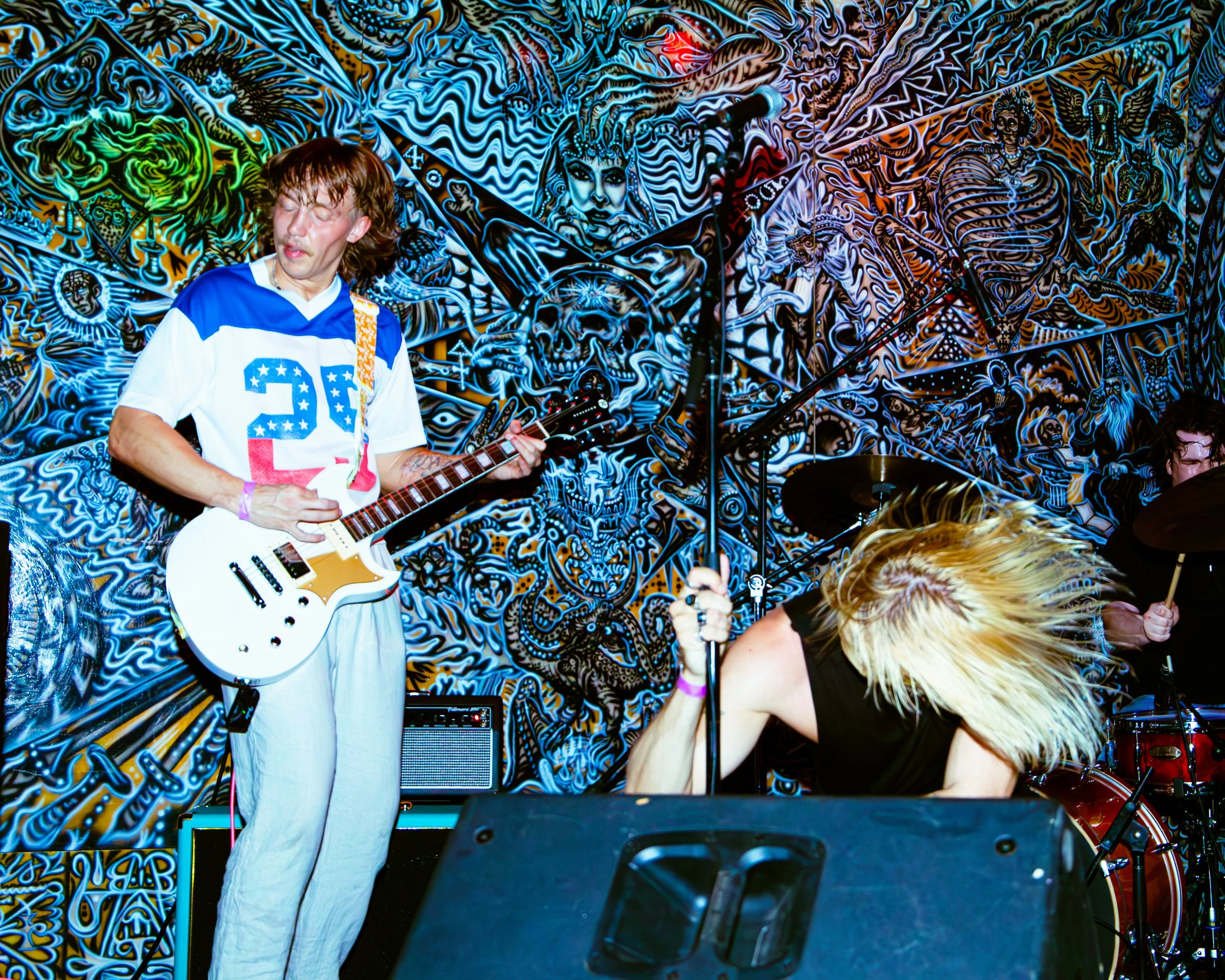 Musicians performing in front of a colorful, psychedelic mural at a concert. One person with long hair and a white jersey playing an electric guitar, another person with blonde hair dancing, and a background featuring intricate, vivid artwork.