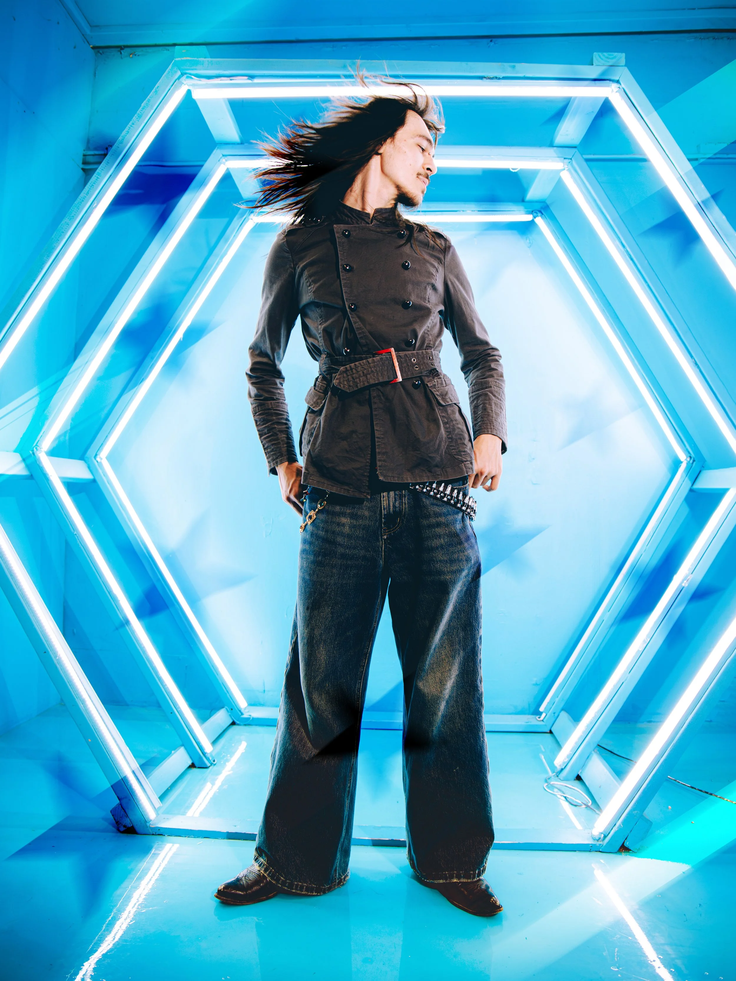 Local Dallas, Texas, DFW Non-traditional Fashion Editorial Creative Portraits Cover Art Photographer Photoshoot. A person with long hair in motion, wearing a dark jacket and wide-leg jeans, standing in a futuristic, neon-lit tunnel with blue and whit