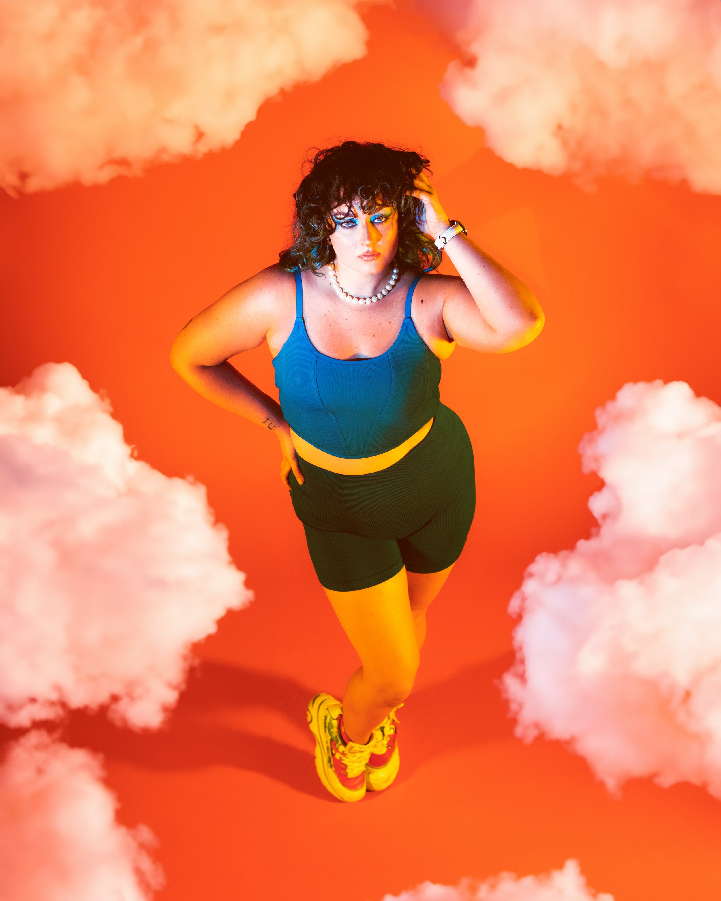 Local Dallas, Texas, DFW Non-traditional Fashion Editorial Creative Portraits Cover Art Photographer Photoshoot. Musician Ravs. Person with curly hair wearing a blue crop top, black shorts, yellow sneakers, a pearl necklace, and a wristwatch posing a