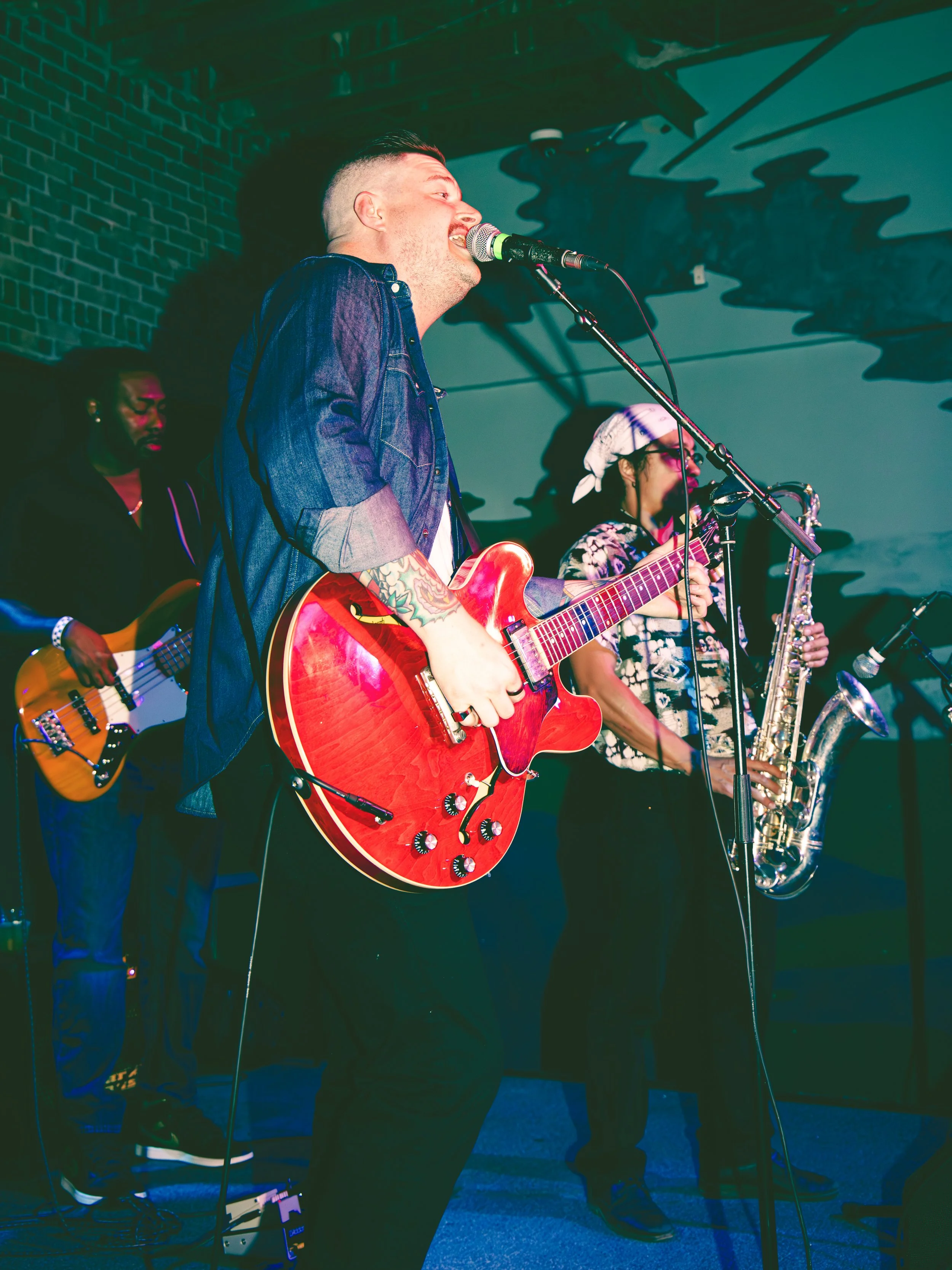 A band performing on stage with a singer playing an acoustic guitar, a bass player, and a saxophone player. The singer is wearing a denim jacket and has tattoos on his arm. The stage has a brick wall background and stage lighting.