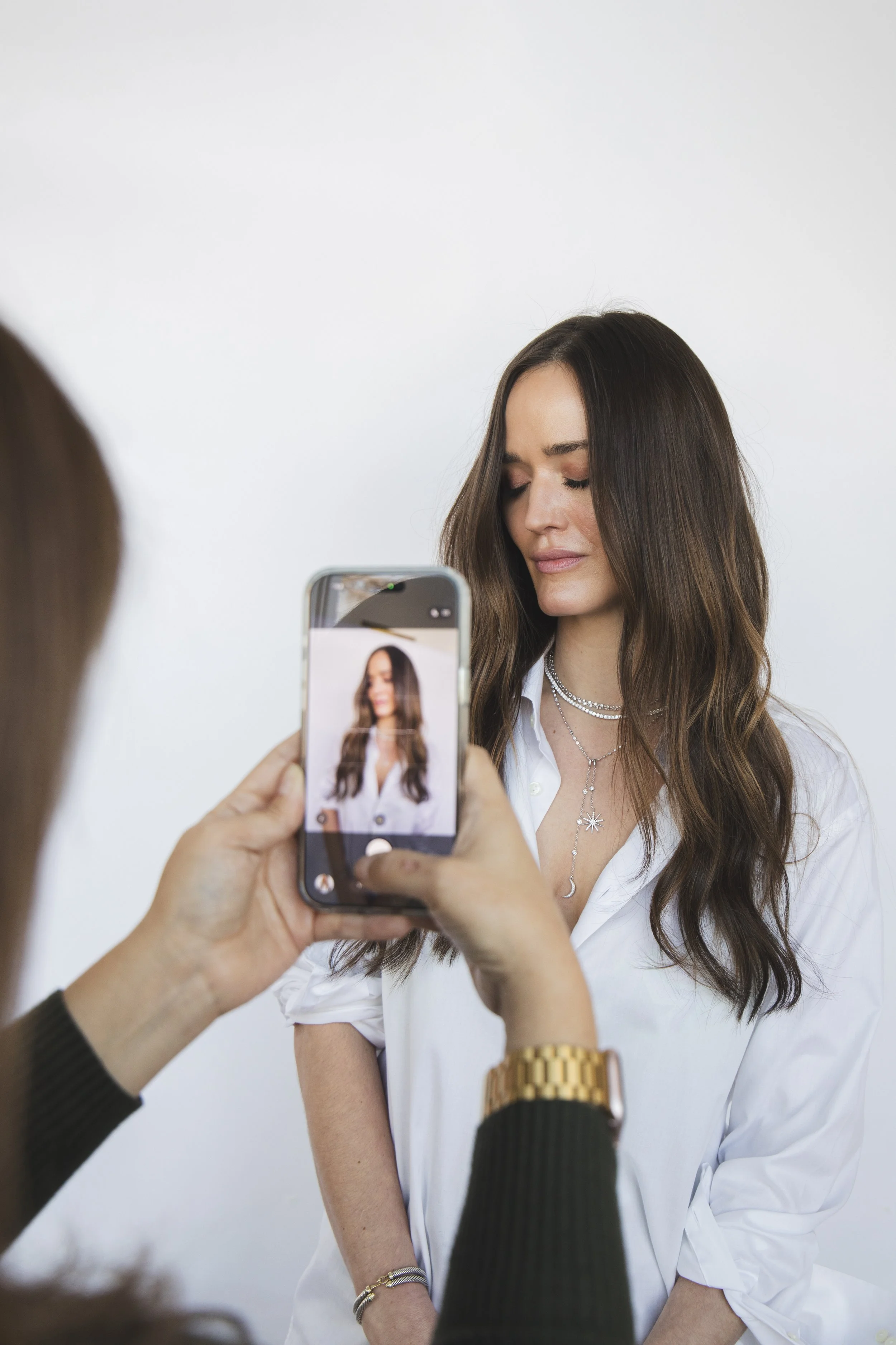 Local Dallas, Texas, DFW Non-traditional Fashion Editorial Creative Portraits Cover Art Photographer Photoshoot. Revel Workshop . A woman with long brown hair, wearing a white shirt and layered necklaces, is being photographed by another person holdi