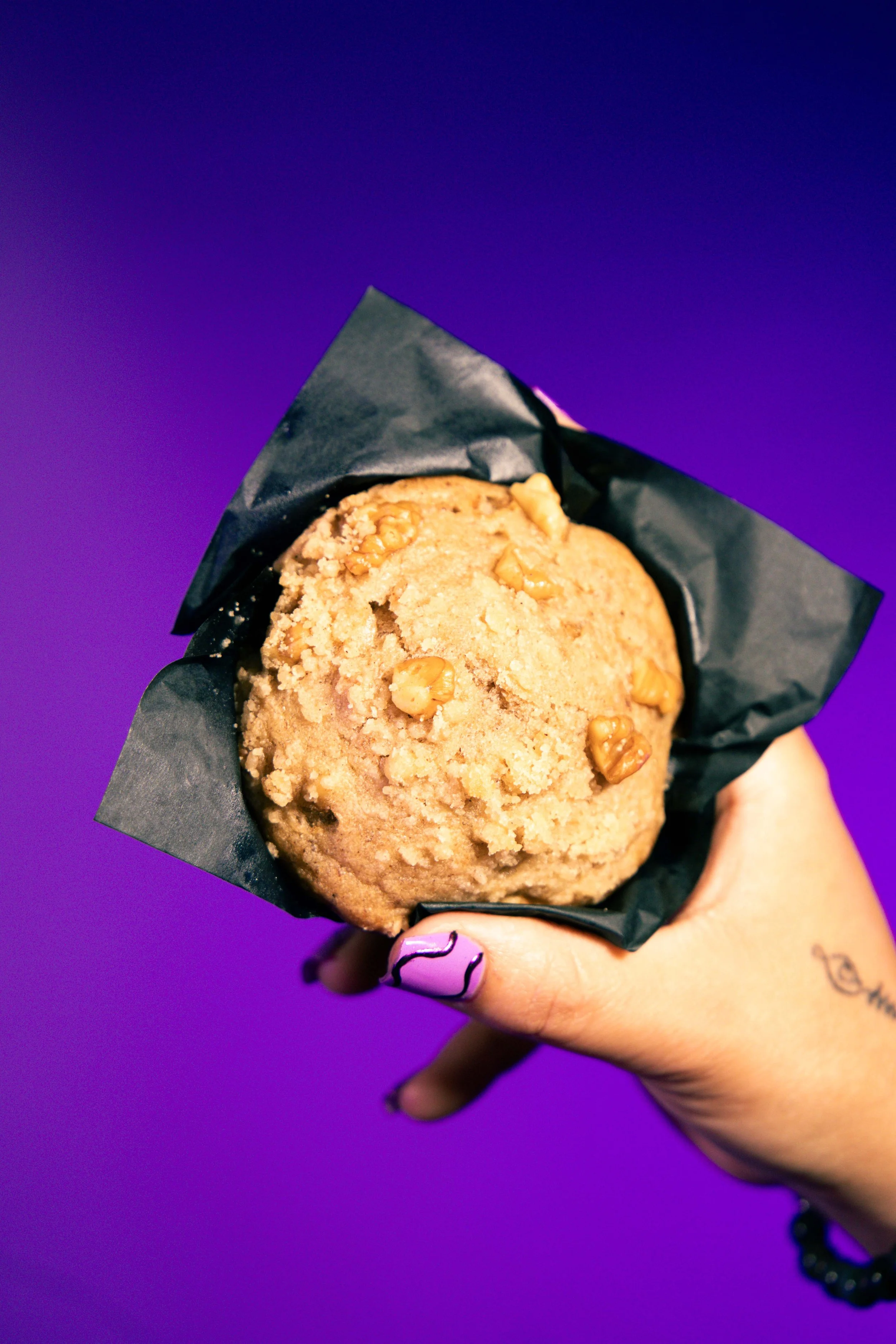Close-up of a cookie with walnuts in black wrapping, held by a hand with painted nails against a purple background.