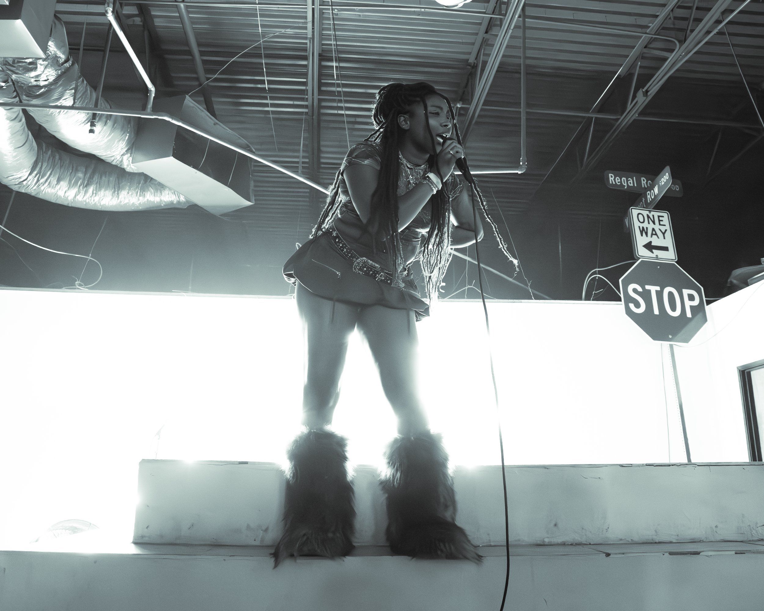 A woman with dreadlocks singing into a microphone at an indoor event, with a sign indicating a street corner at Regal Road and Row Avenue. The photo is in black and white with high contrast.