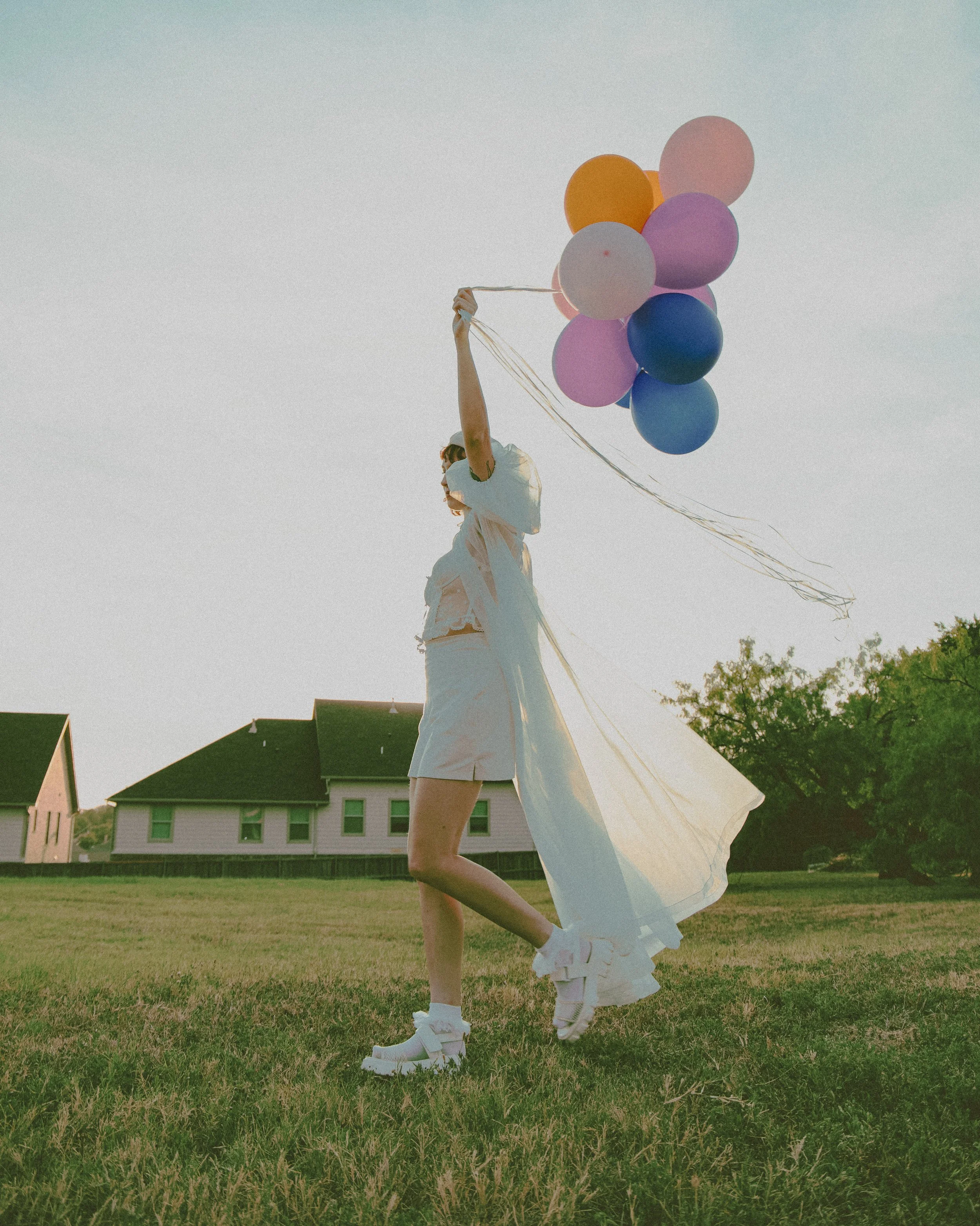 Person in a white dress and roller skates holding a bunch of colorful balloons outdoors during sunset.