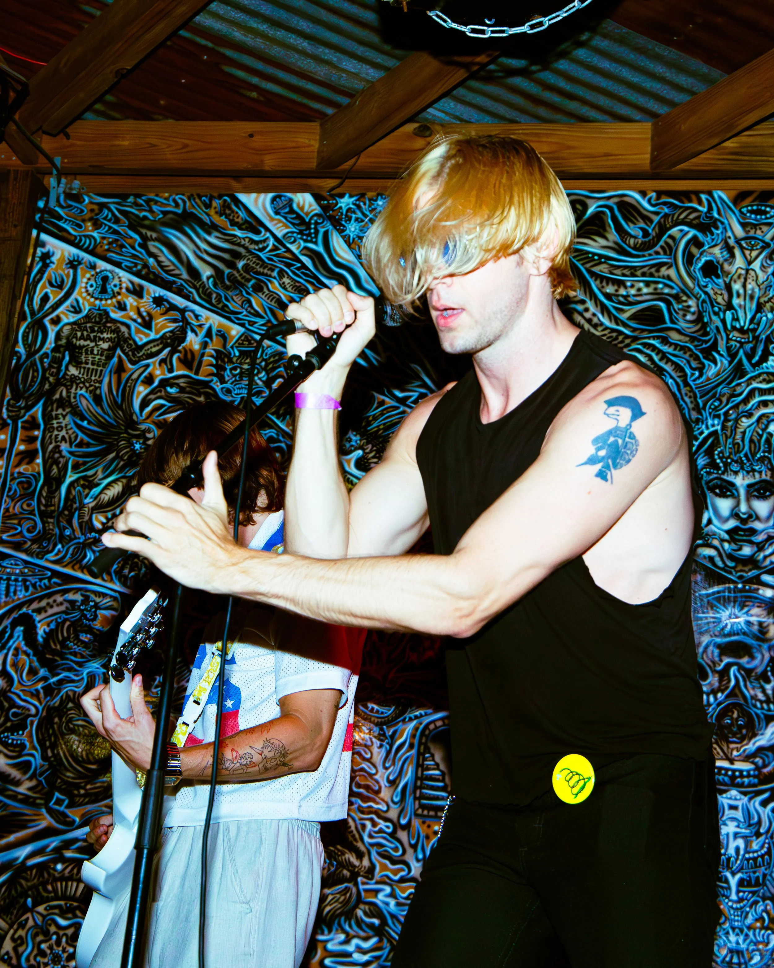 A musician with long blond hair, wearing a black sleeveless shirt, singing into a microphone, with tattoos on his arm, and a yellow smiley face sticker on his pants. Behind him is a person wearing a white shirt with a colorful pattern, holding a guit