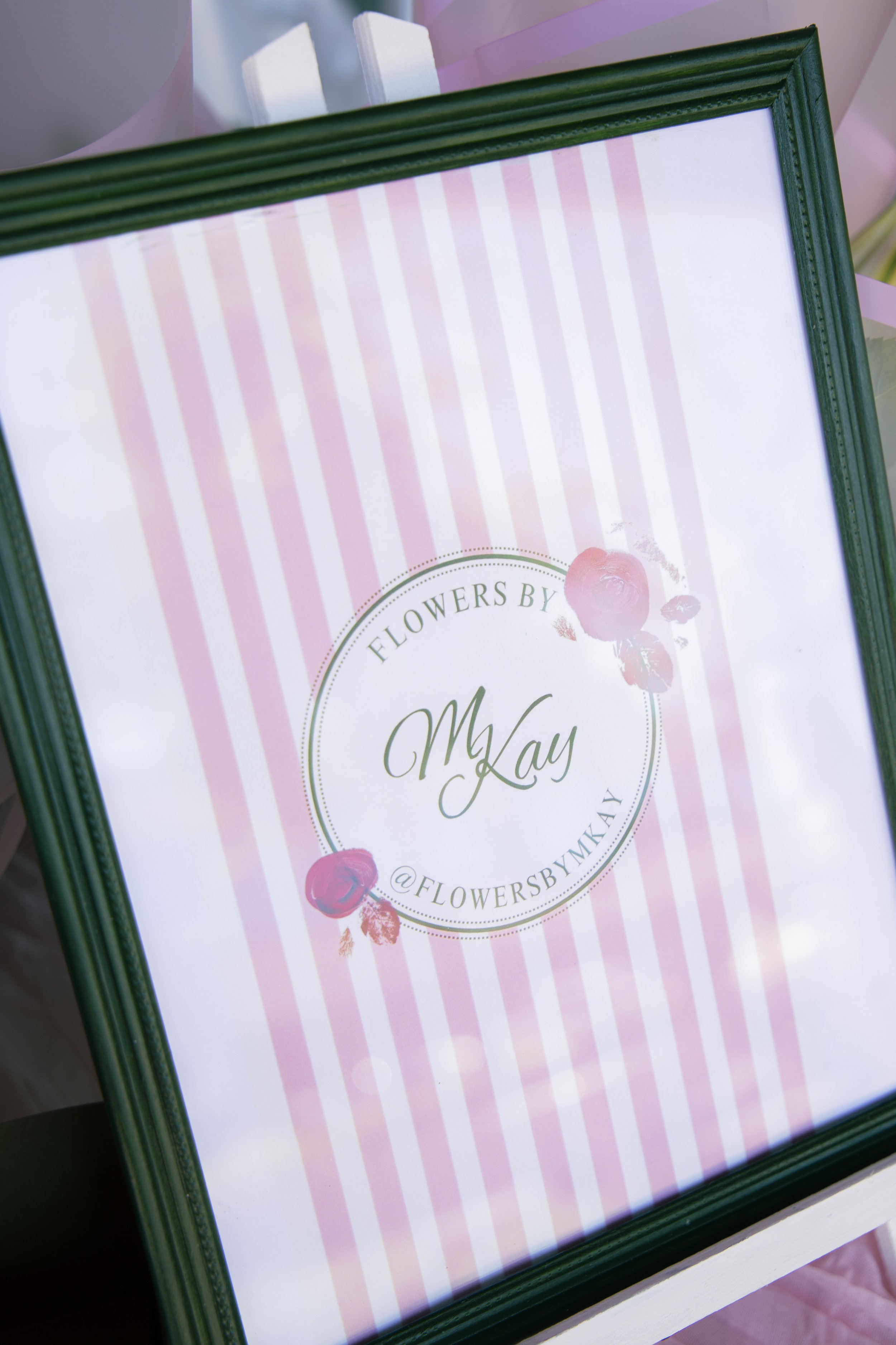 A framed print with pink and white vertical stripes and a circular design in the center with text that reads 'FLOWERS BY' and 'May'. There are pink roses or rose illustrations around the circle. The frame is dark green, and the print appears to be re