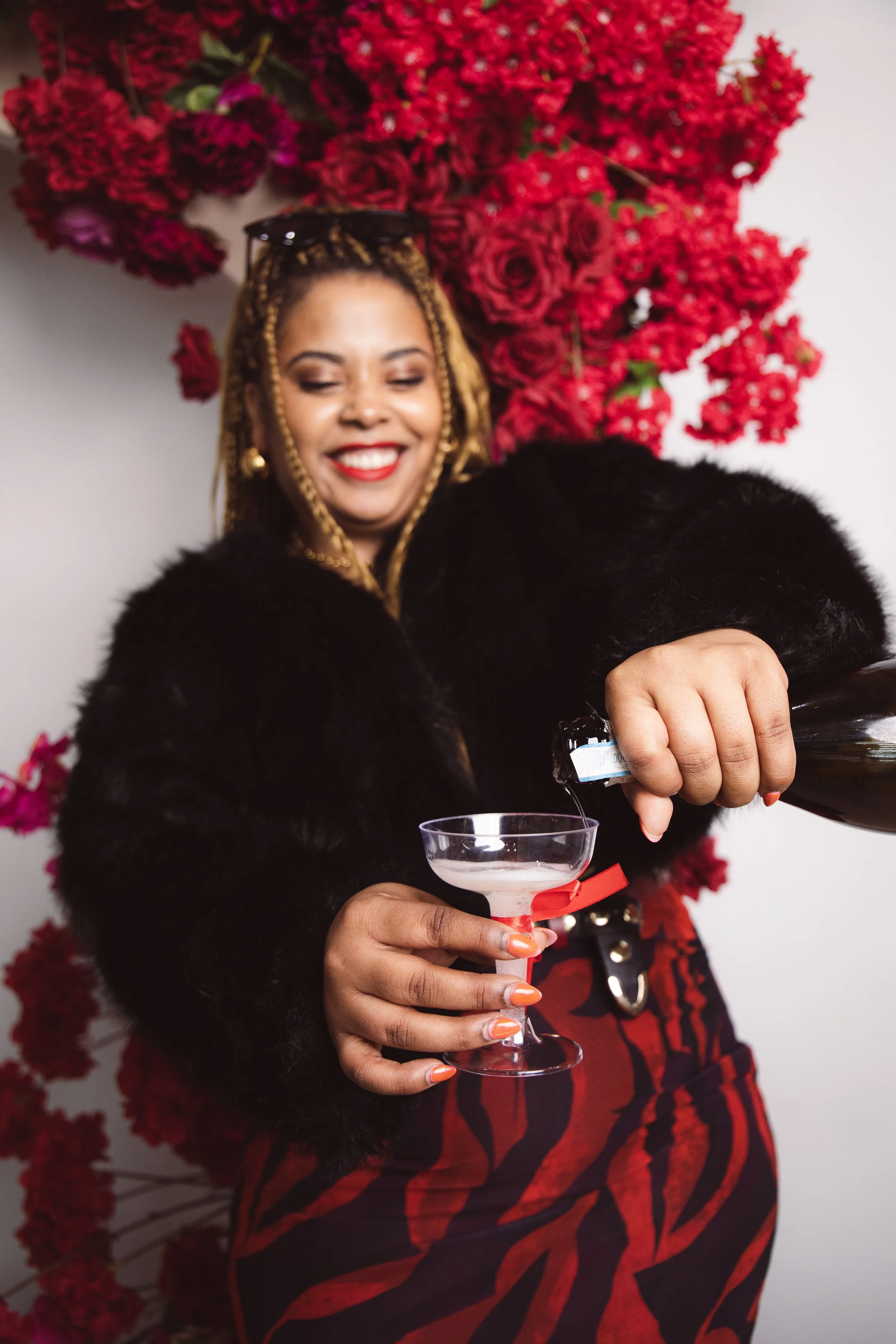 A woman with blonde braids wearing a black fur coat and red lipstick is pouring a drink into a martini glass. She is smiling and standing in front of a backdrop of red flowers. She has sunglasses on her head and is wearing gold earrings.