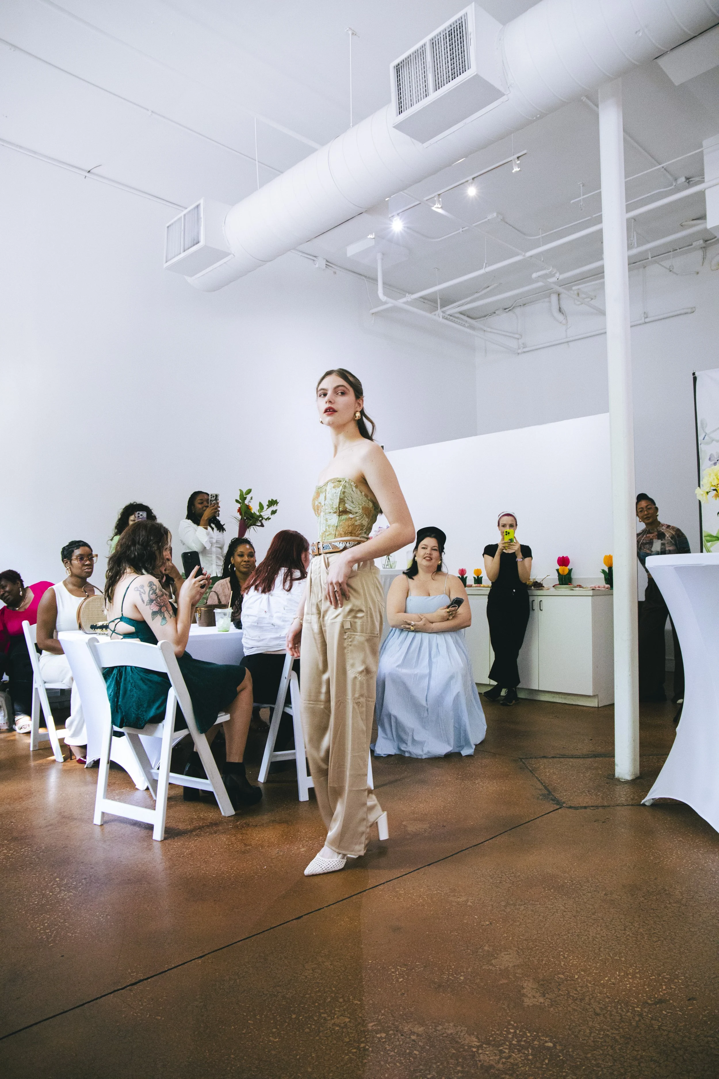 A woman in beige wide-leg pants and a strapless floral top walking in a room with white walls and ceiling, surrounded by seated women, some taking photos, some smiling, with tulips on the counter to the right.