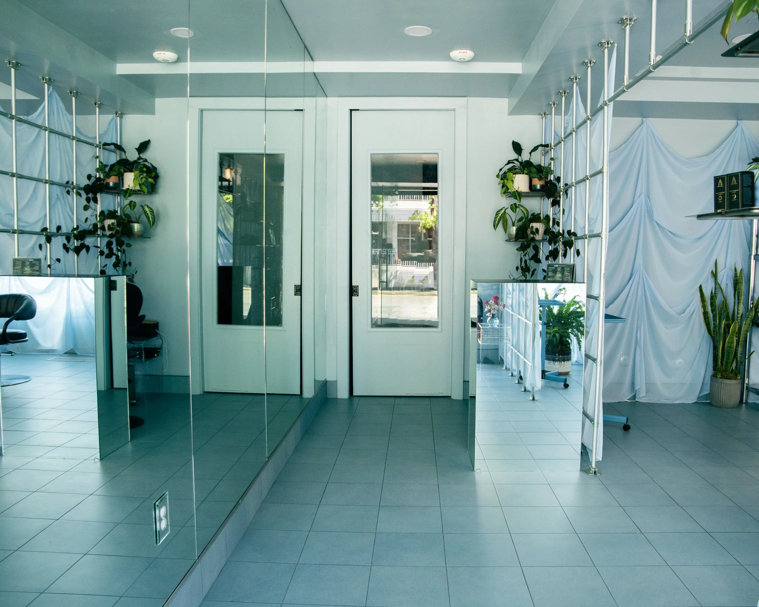 Interior view of a modern building with glass doors, white walls, plants in pots, white curtains, and a reflective mirror wall, with ceiling lights and tiled floor.