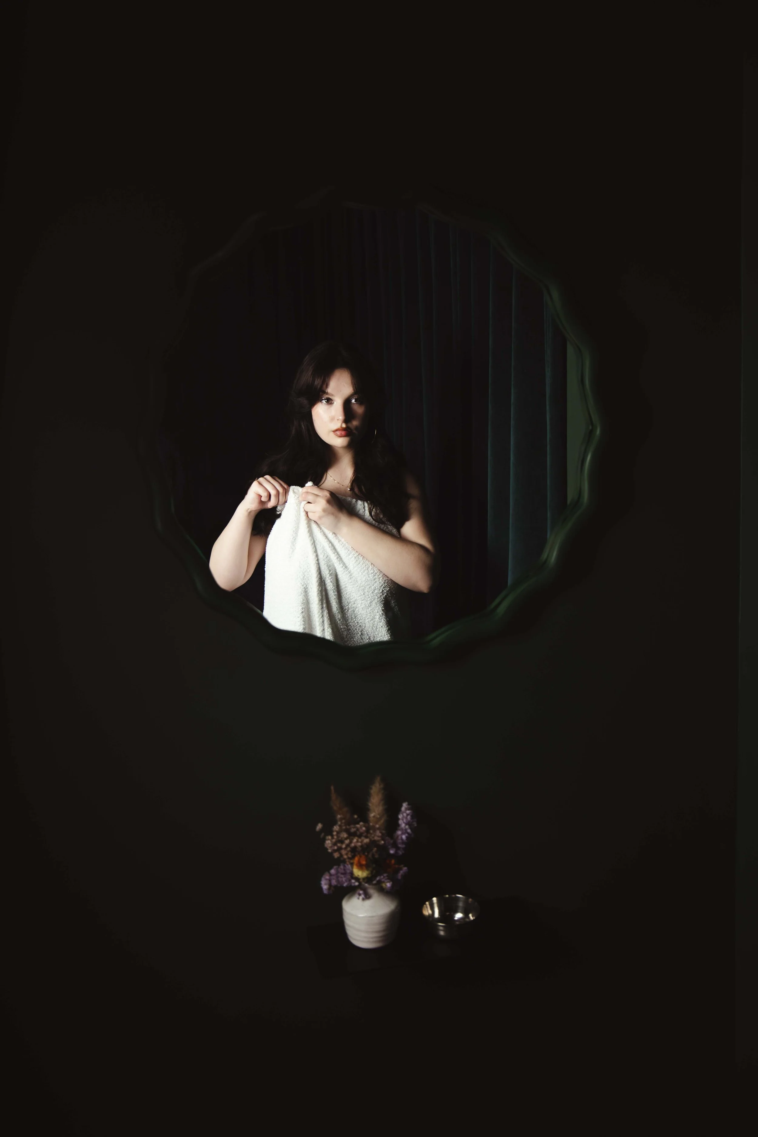 A woman with dark hair and red lipstick looking into a mirror holding a white towel wrapped around her body. The mirror has a wavy green border and reflects a dark background with vertical lines. There is a table below the mirror with a white vase co