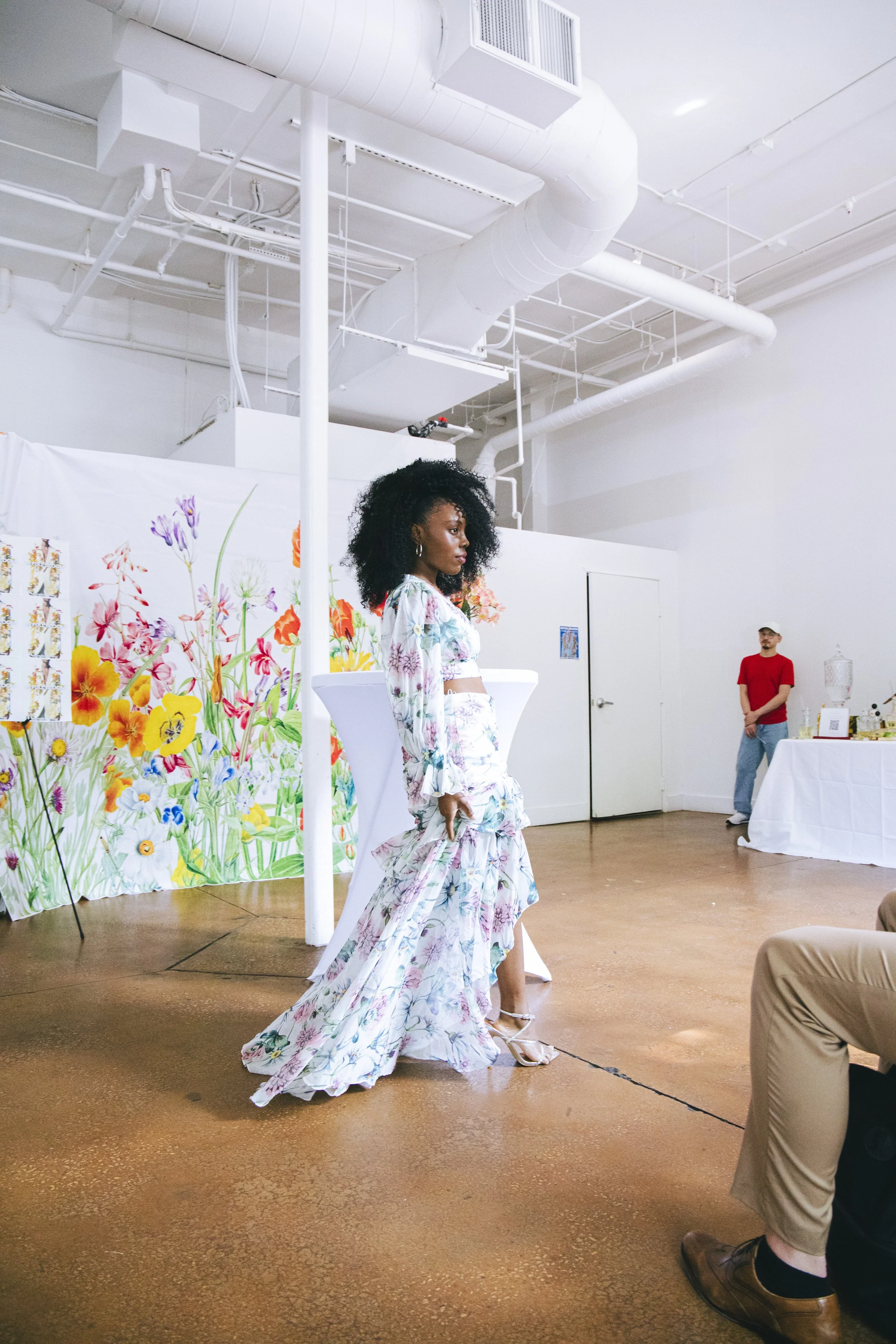 A woman with curly black hair wearing a flowy, floral dress standing in a gallery or art space. Behind her is a colorful floral artwork display. There are other people in the background, including a man in a red shirt and beige cap standing near a ta