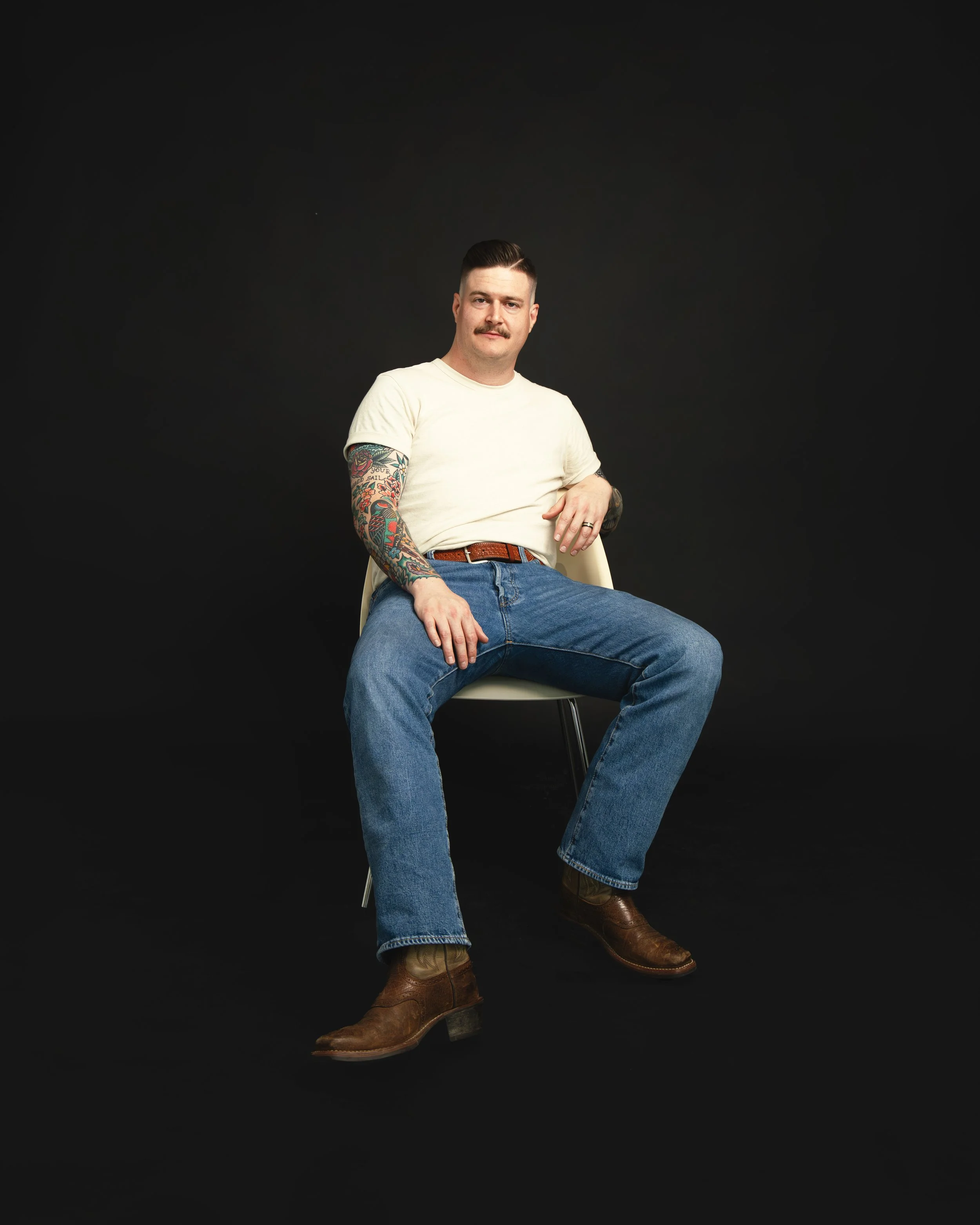 Local Dallas, Texas, DFW Non-traditional Fashion Editorial Creative Portraits Cover Art Photographer Photoshoot. Musician Caleb Rains Band . A man with a mustache, tattoos on his left arm, wearing a white T-shirt, blue jeans, and brown cowboy boots, 
