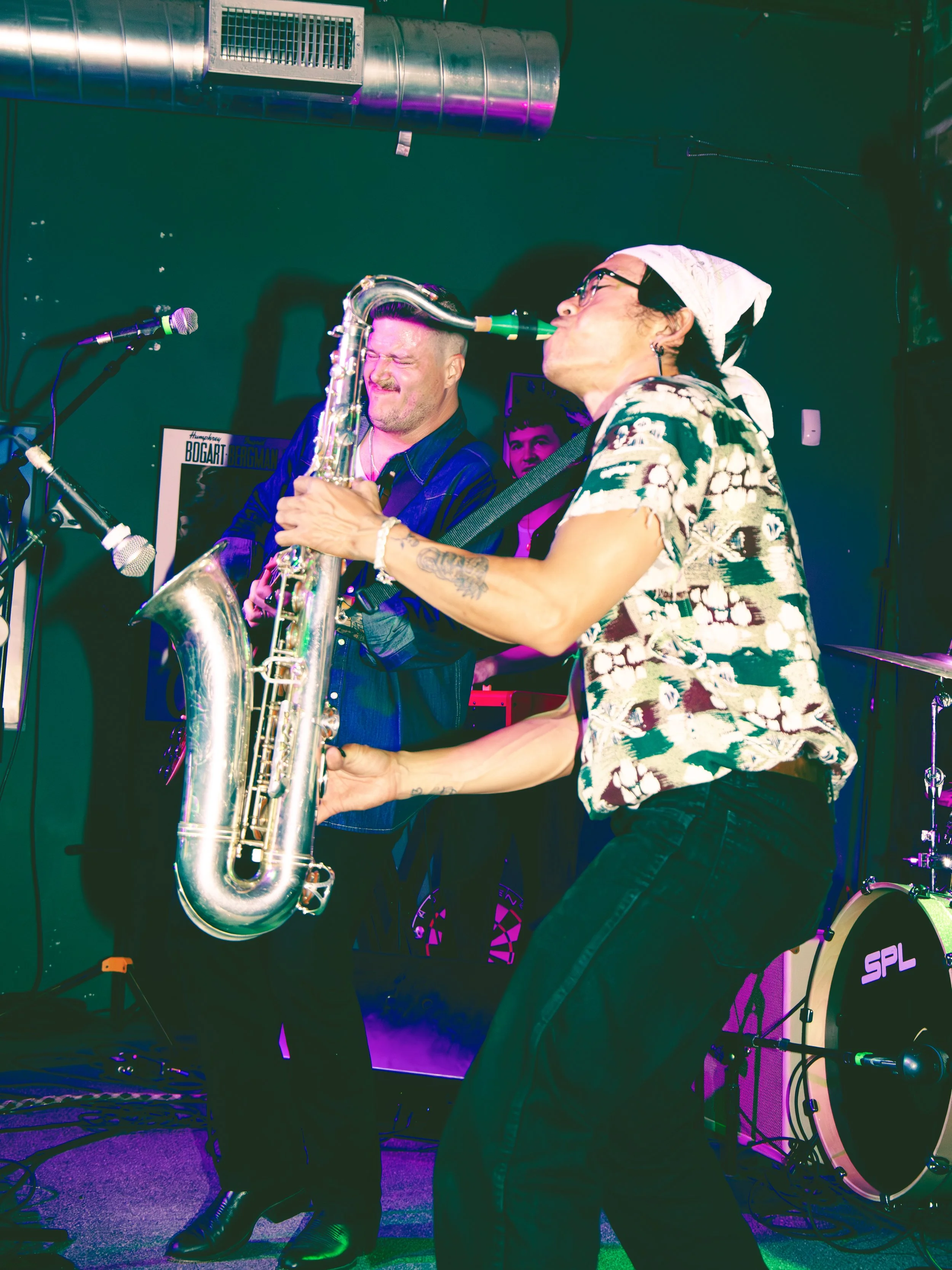 Two musicians performing on stage with one playing a saxophone and the other singing into a microphone, with colorful stage lighting and musical equipment around them.