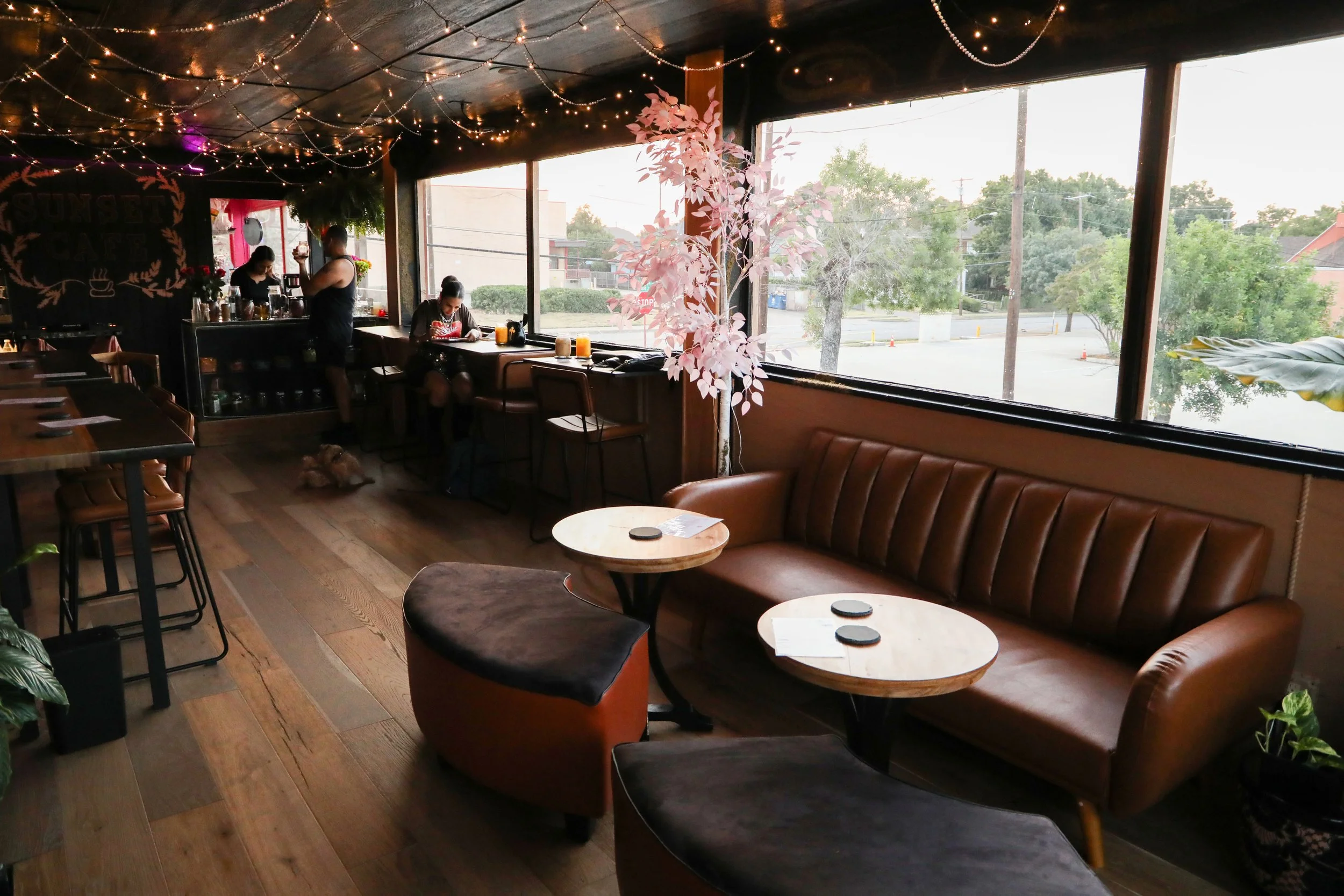 Interior of a cozy cafe with large windows showing trees and street outside, string lights on the ceiling, a pink fake tree near the window, and a barista behind the counter preparing drinks. There are empty tables and chairs, and a person sitting at